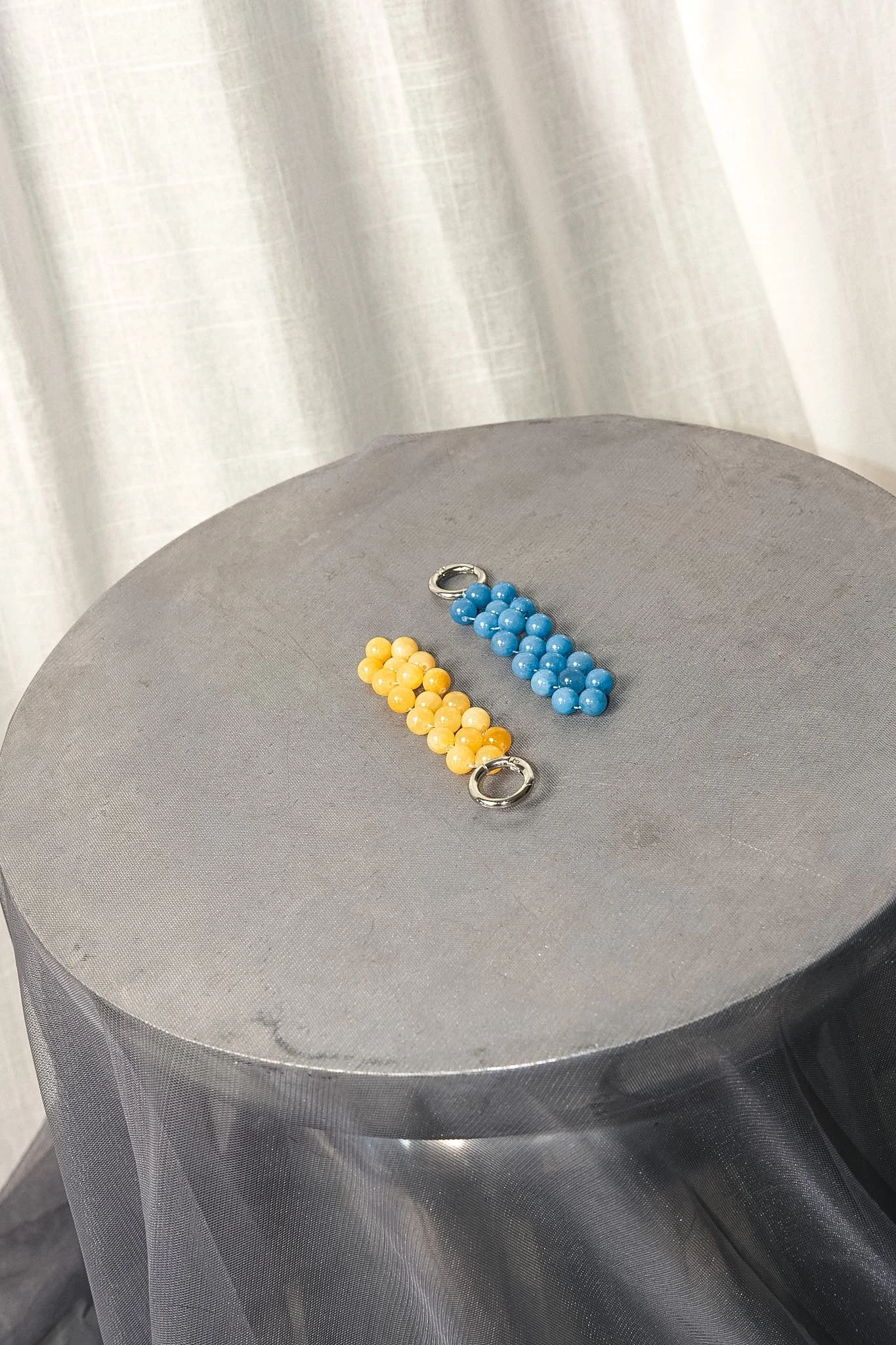 Two beaded bag charms, one yellow and one blue, with silver clasps, resting on a round gray surface.