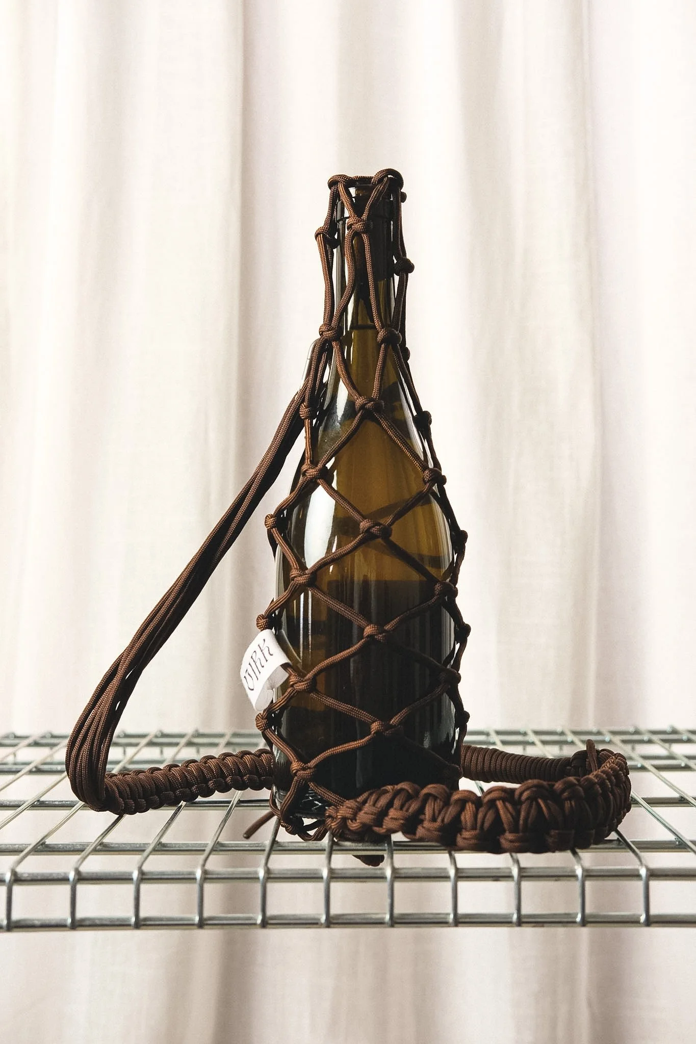 A brown glass bottle inside a brown netted paracord bottle holder with a matching bottle leash, placed on a metal wire shelf against a white curtain background.