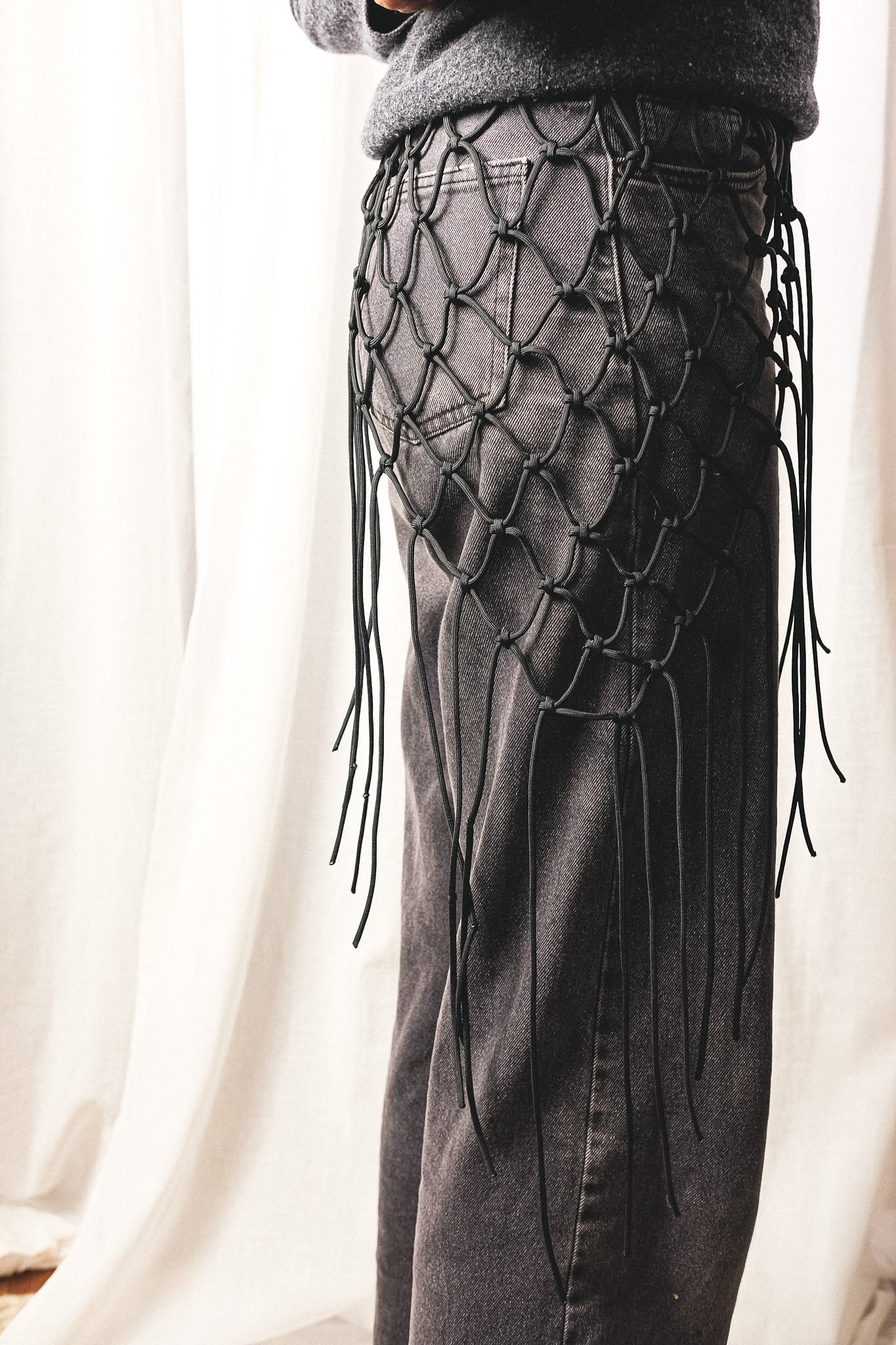 Person wearing a black macrame accessory over dark jeans, with a gray oversized sweater.