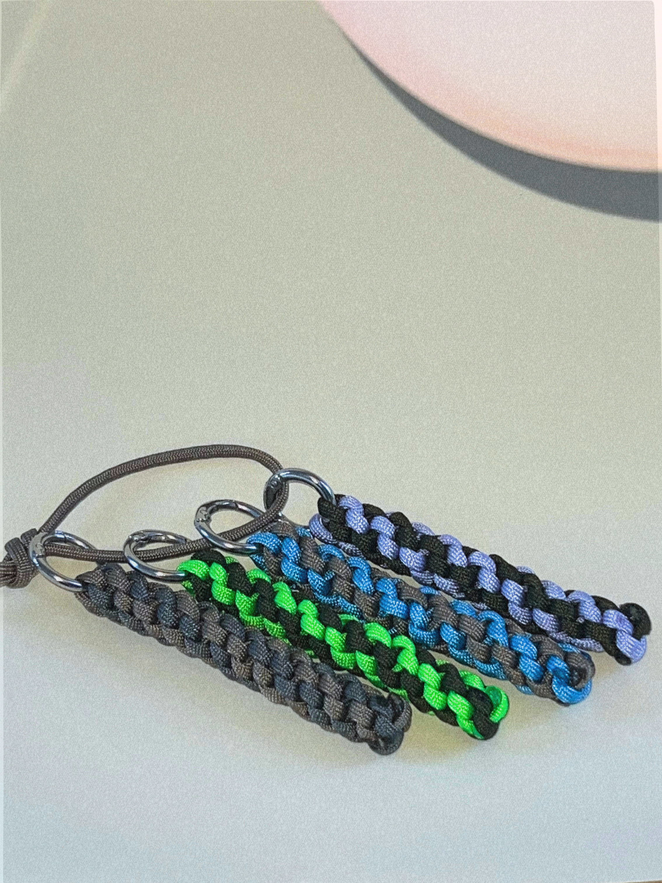 A collection of colorful paracord keyrings, featuring gray, black, green, blue, and purple cords, with some having metallic rings attached.