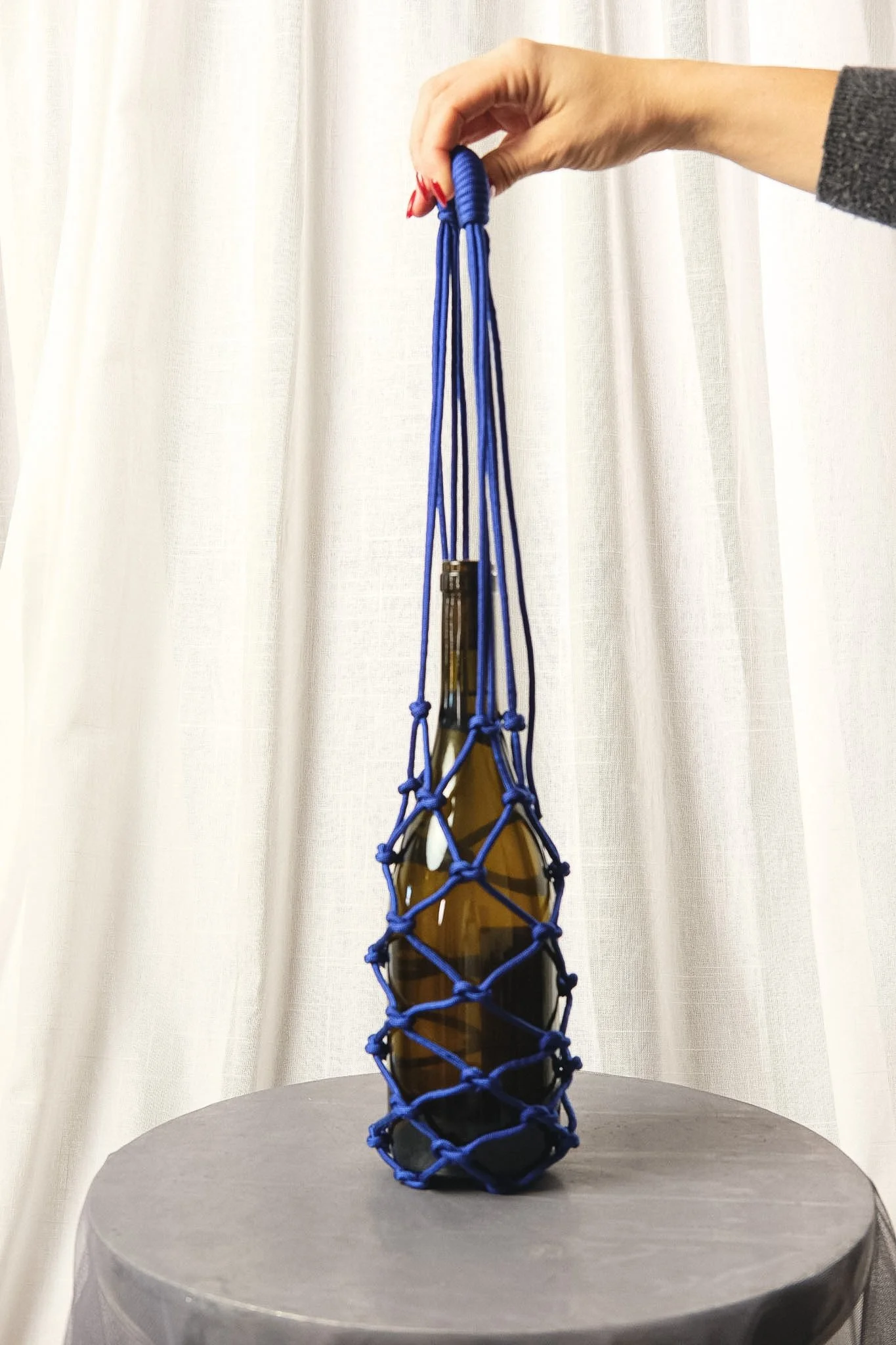 Hand holding a blue rope net surrounding a brown glass bottle on a black round table with a white curtain background.