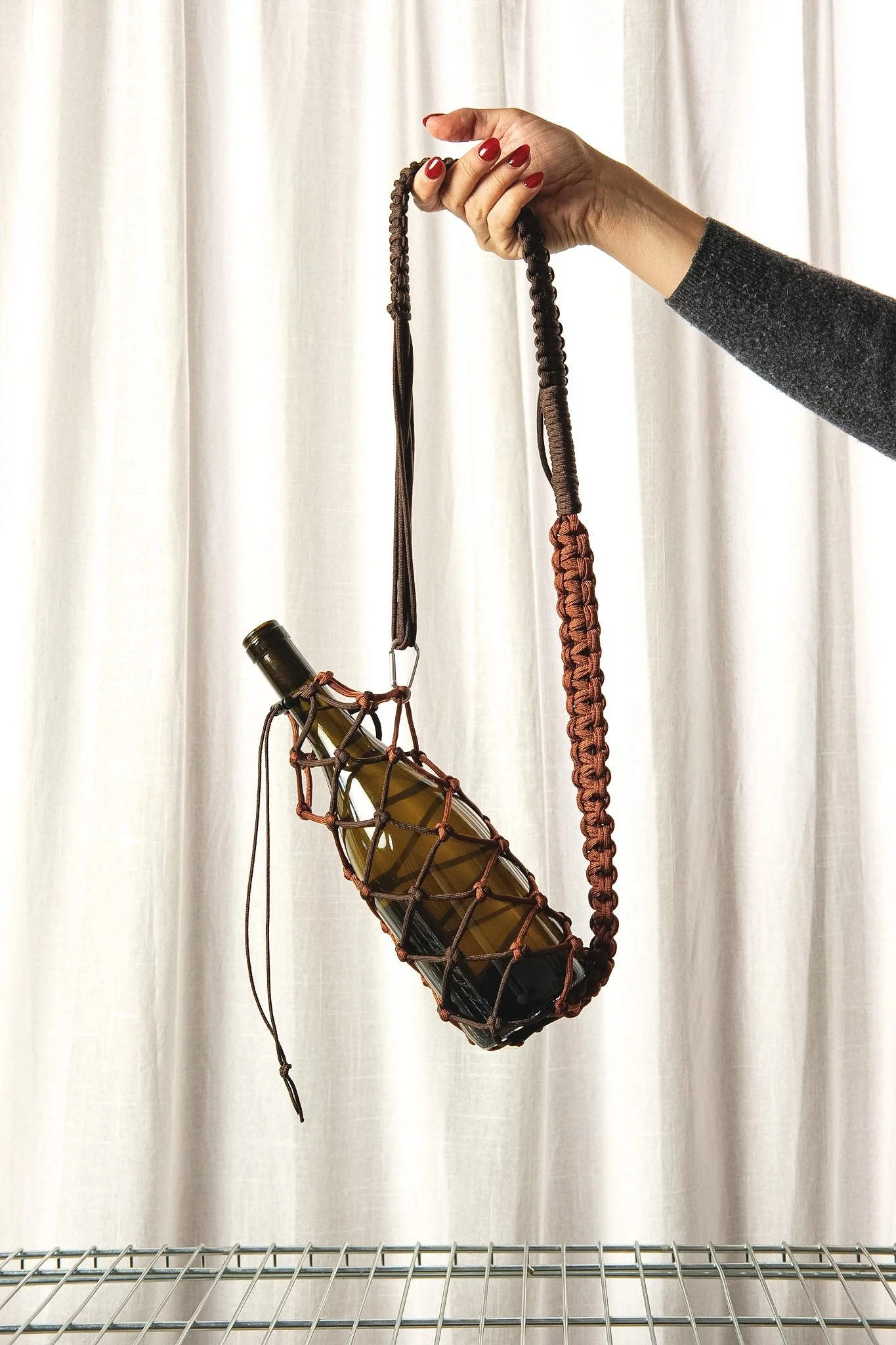 macrame wine holder with a bottle of wine inside against a white curtain background.