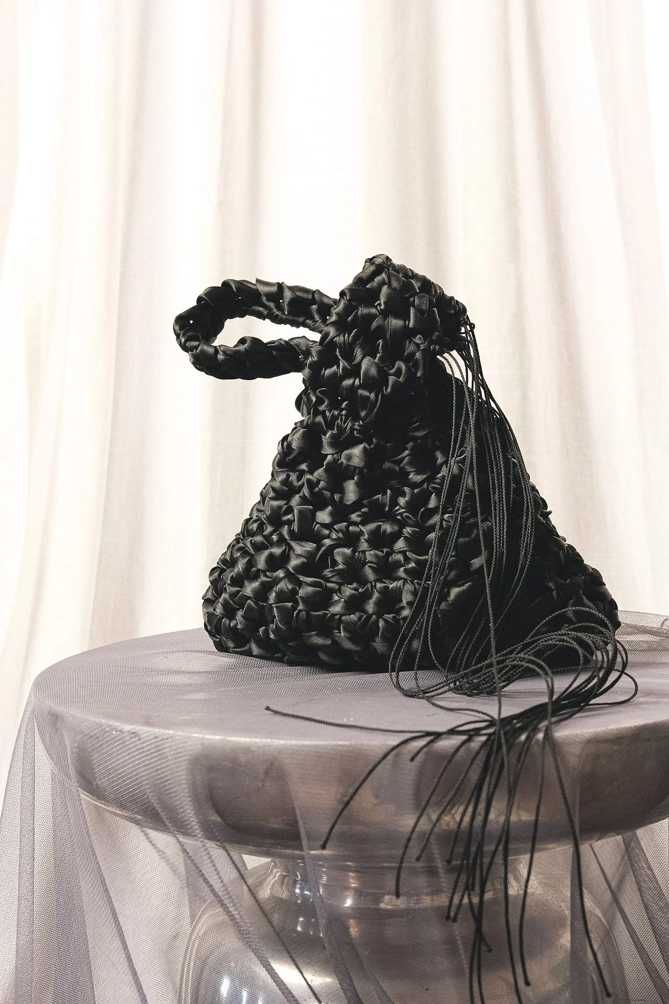 Black woven bag with hanging black cords placed on a round table covered with a sheer white tablecloth, with white curtains in the background.