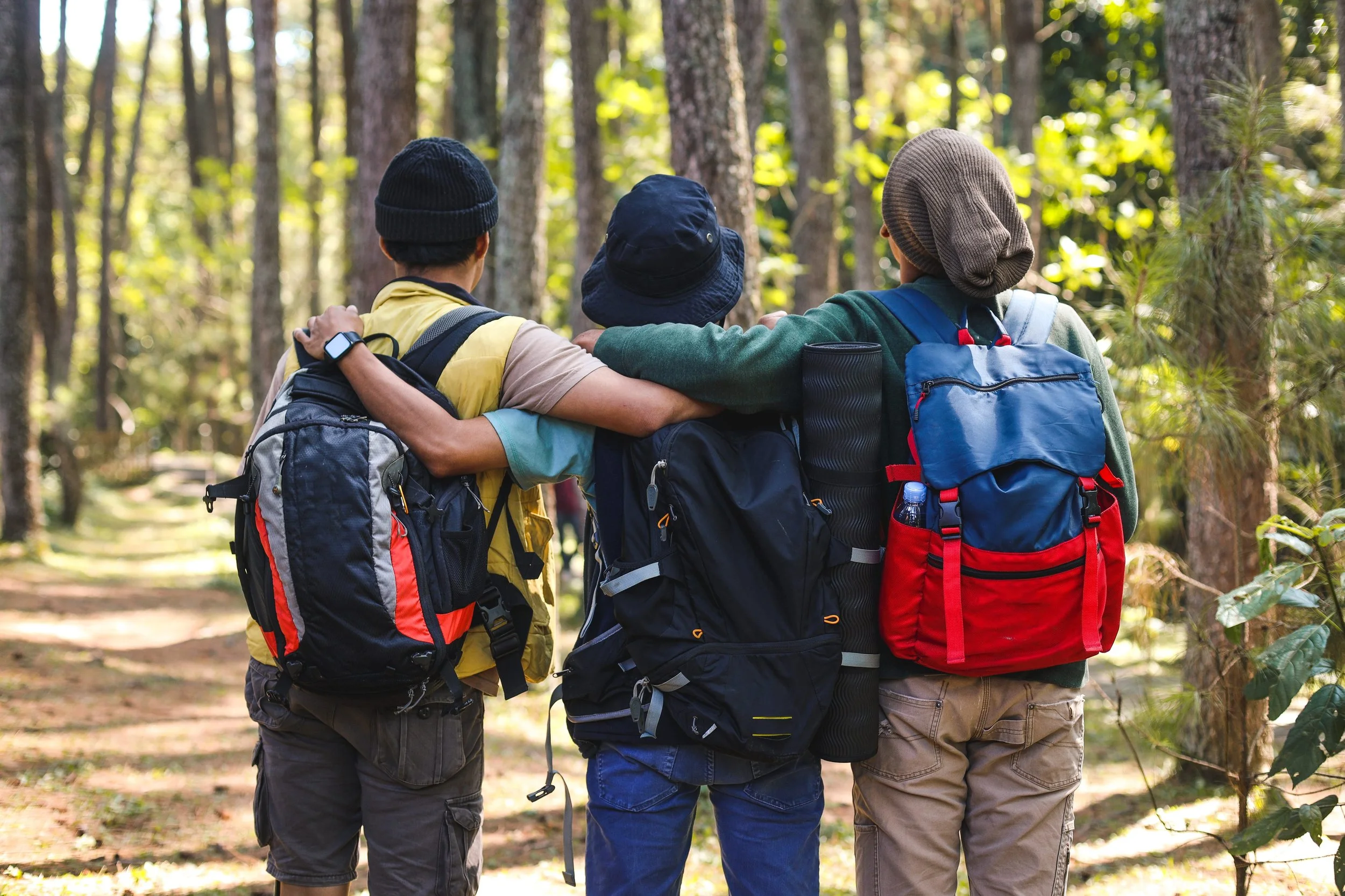 Three hikers with backpacks walking arm-in-arm in the woods, showing friendship, support, and belonging outdoors.
