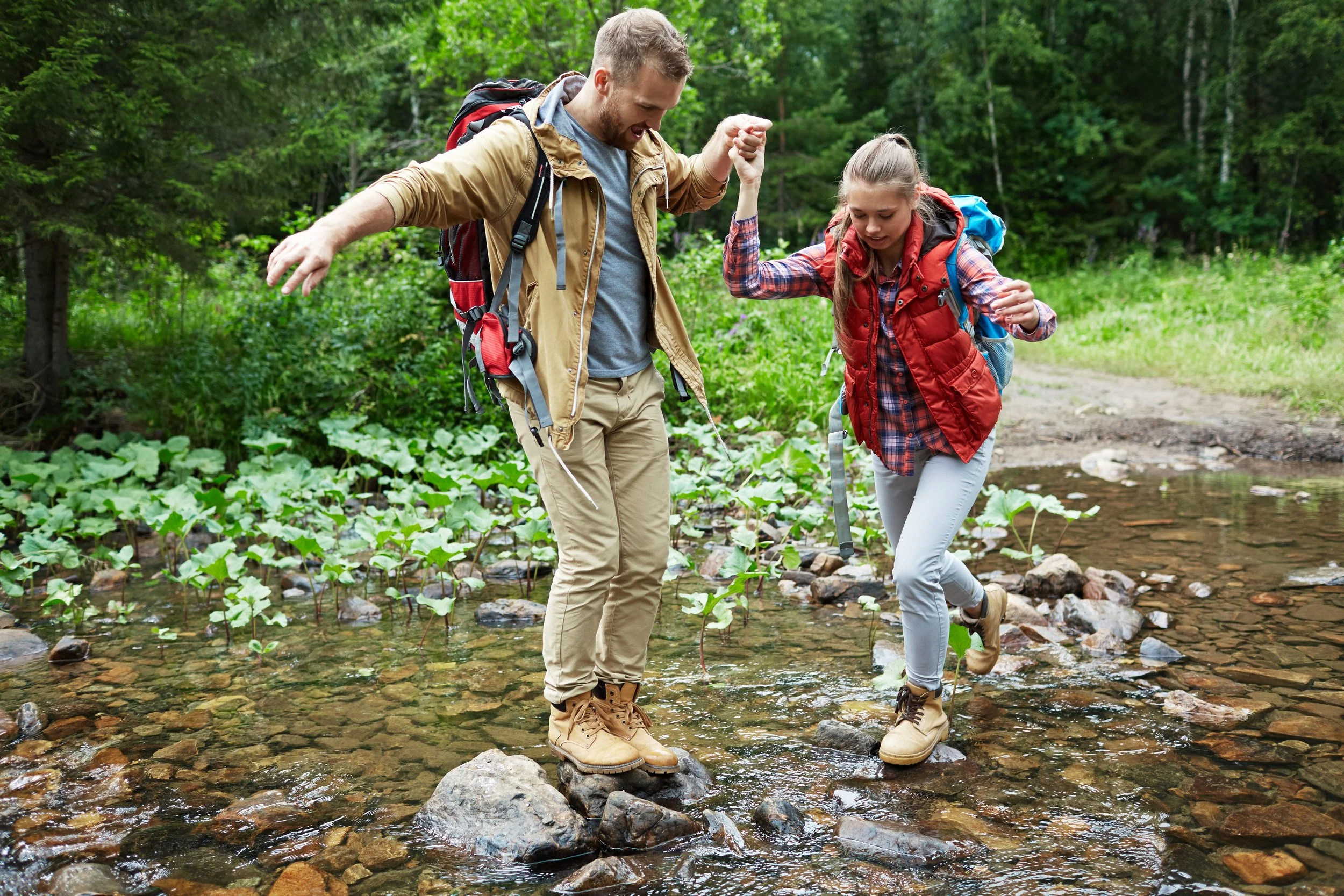 Two young hikers crossing a shallow forest creek on stepping stones while holding hands, representing Sierra Summit Foundation’s mission to build confidence, support, and outdoor access.