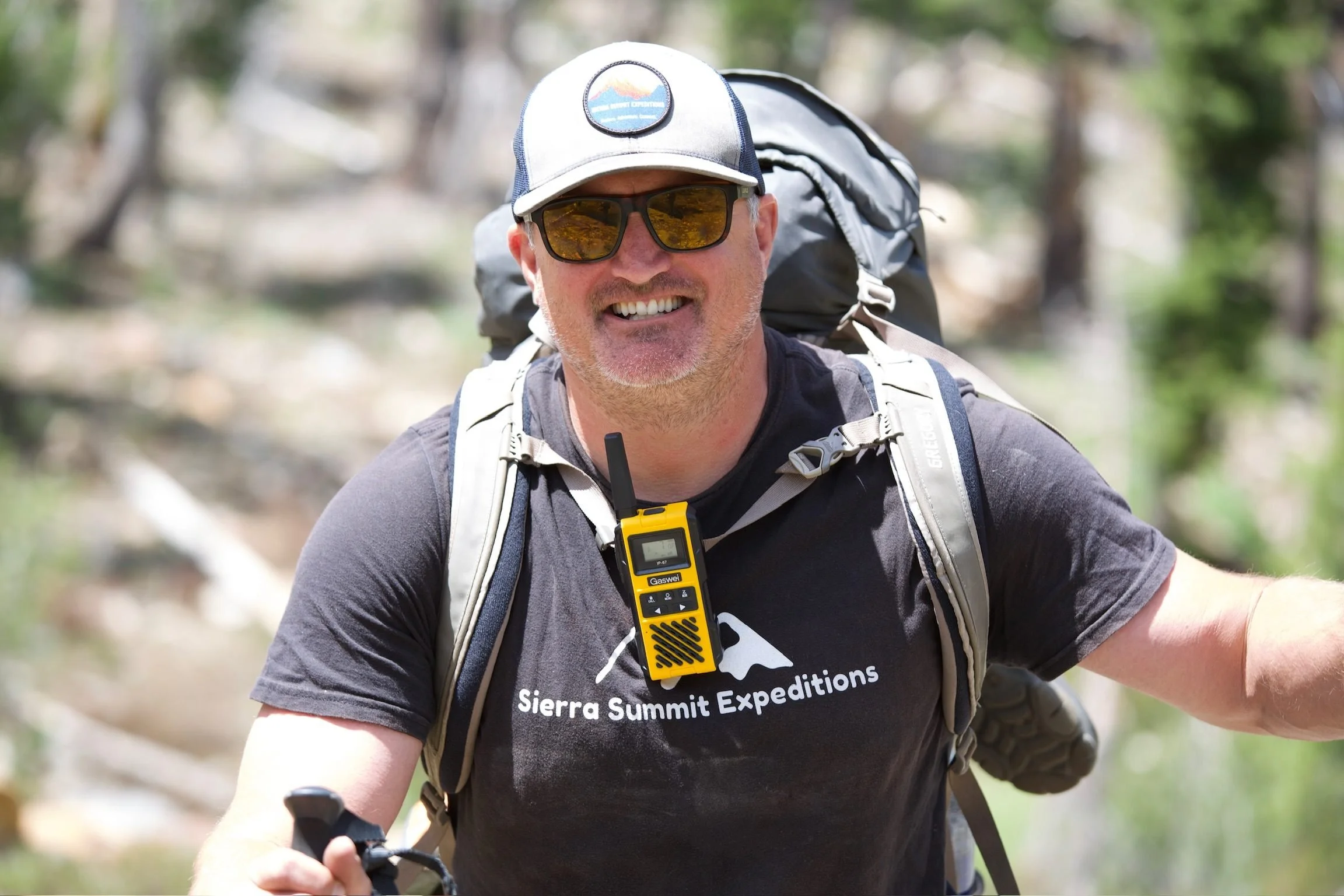 Sierra Summit Expeditions guide smiling on trail wearing a backpack and carrying safety gear, supporting Sierra Summit Foundation programs with trained outdoor leadership.