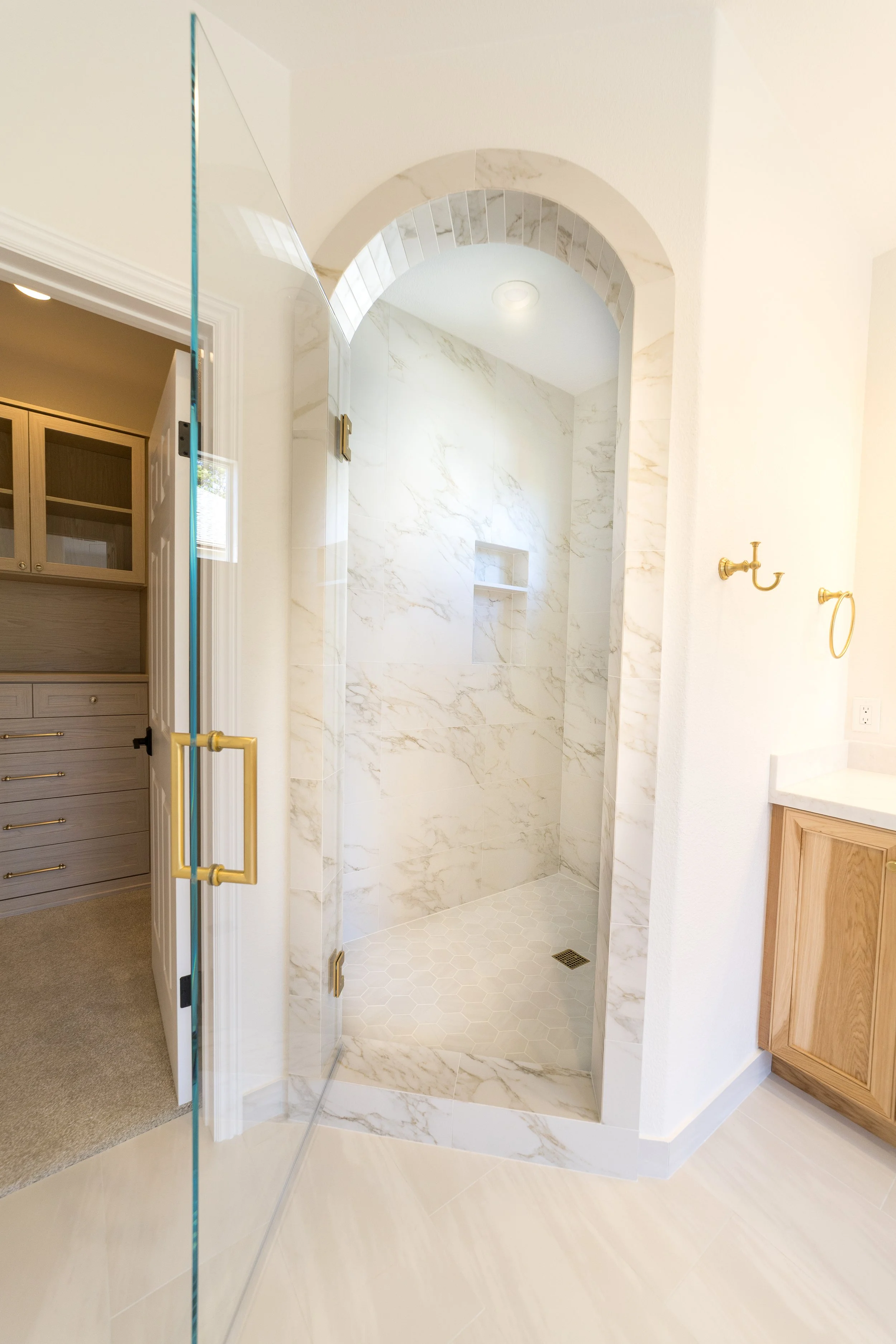 Bathroom with a glass door shower enclosure, white marble walls, a built-in shelf, and a beige tiled floor.
