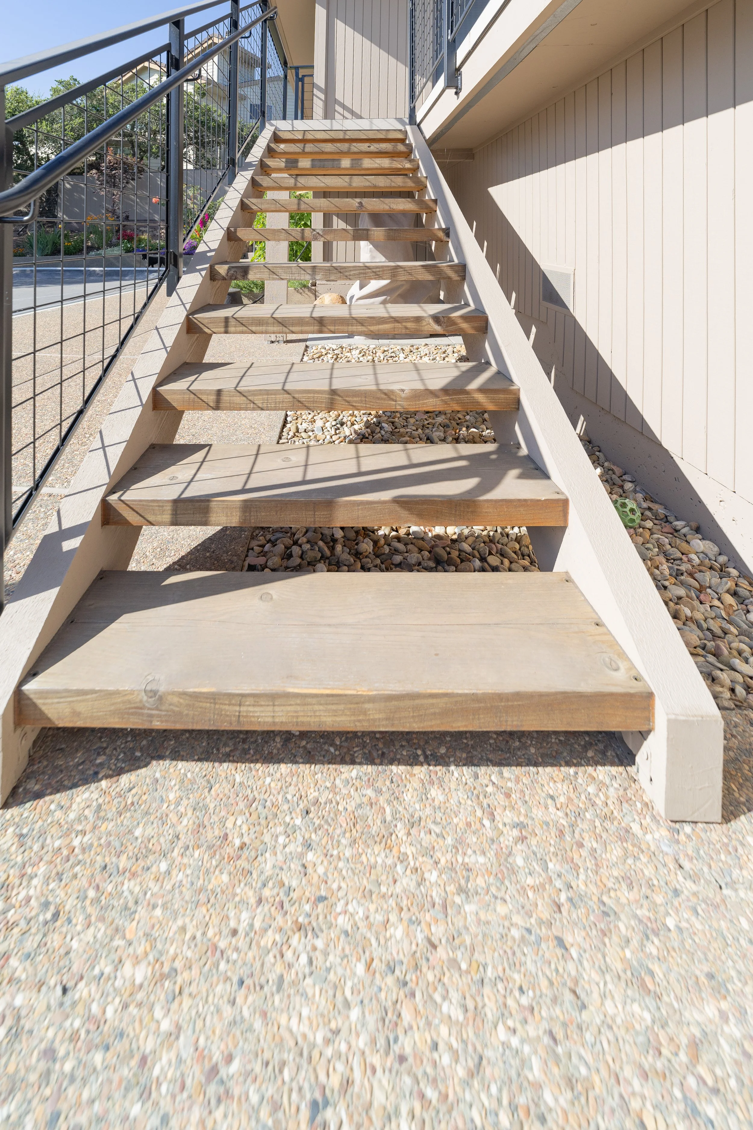 Outdoor staircase with wooden steps, metal railings, leading up to a second-floor patio area, with a dog lying under the stairs in a gravel strip.