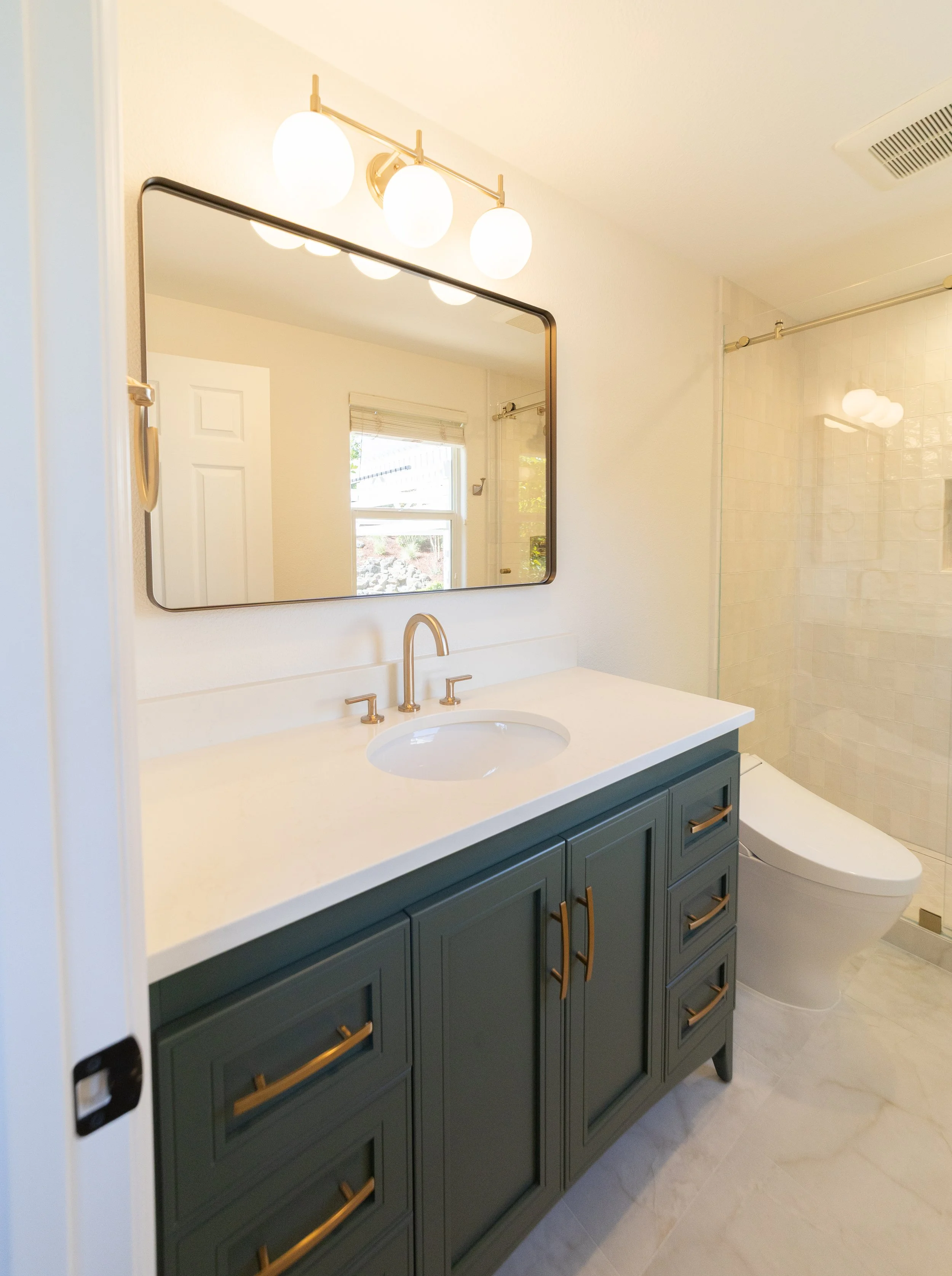 Bathroom with a green vanity, large mirror, white countertop, modern faucet, and a toilet, with a window and a shower area in the background.