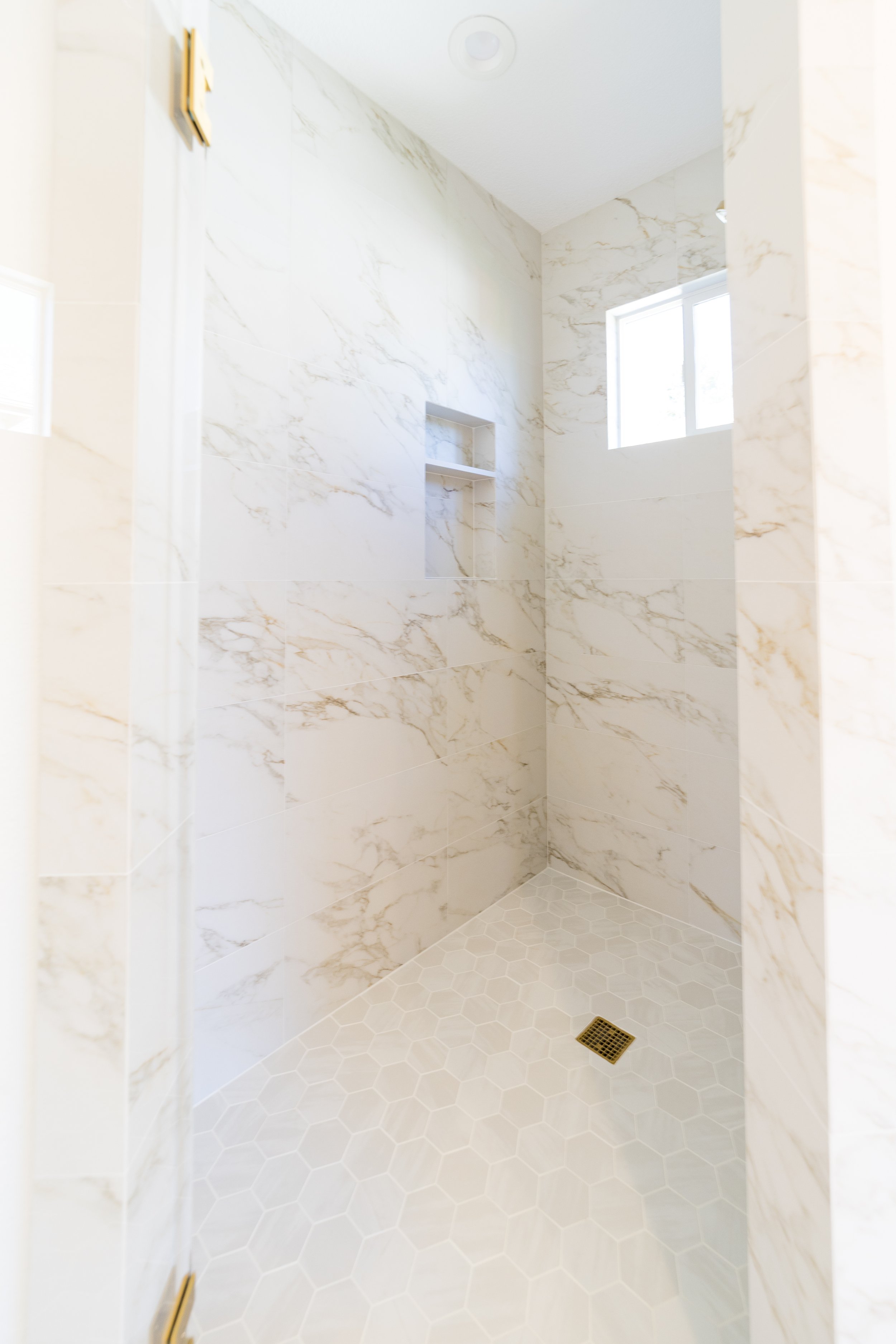 Clean, modern shower with white marble walls, small hexagonal white floor tiles, a small window, built-in soap niches, and a floor drain.