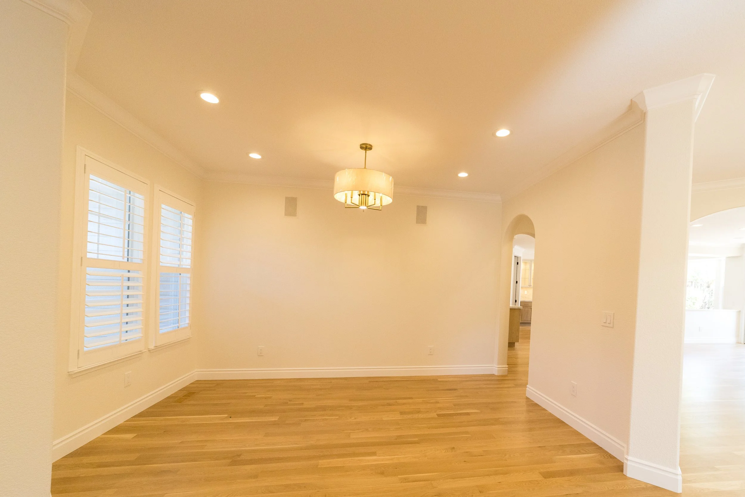 Empty room with wood flooring, white walls, three windows with shutters, and a chandelier hanging from the ceiling.
