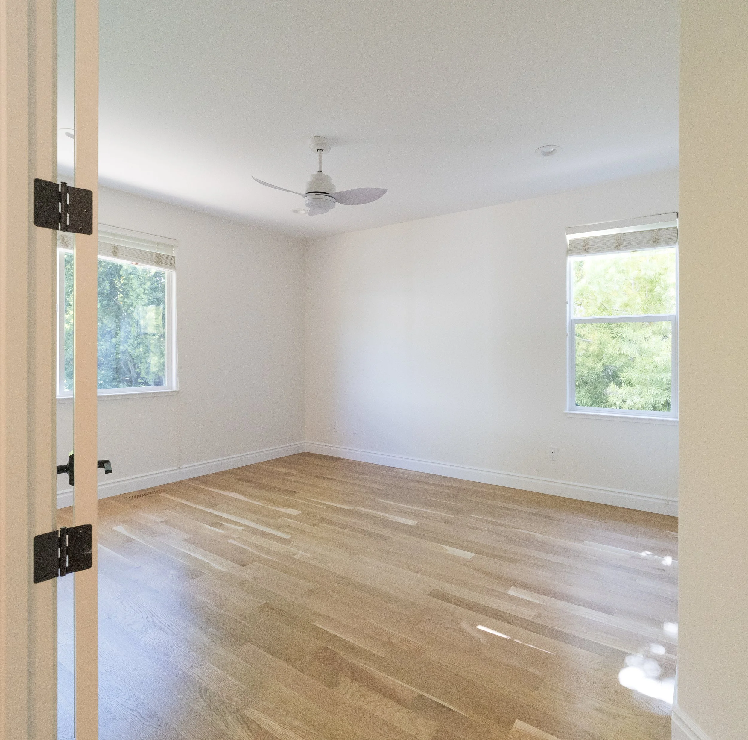 Empty room with hardwood floors, white walls, two windows with blinds, a ceiling fan, and a door frame.