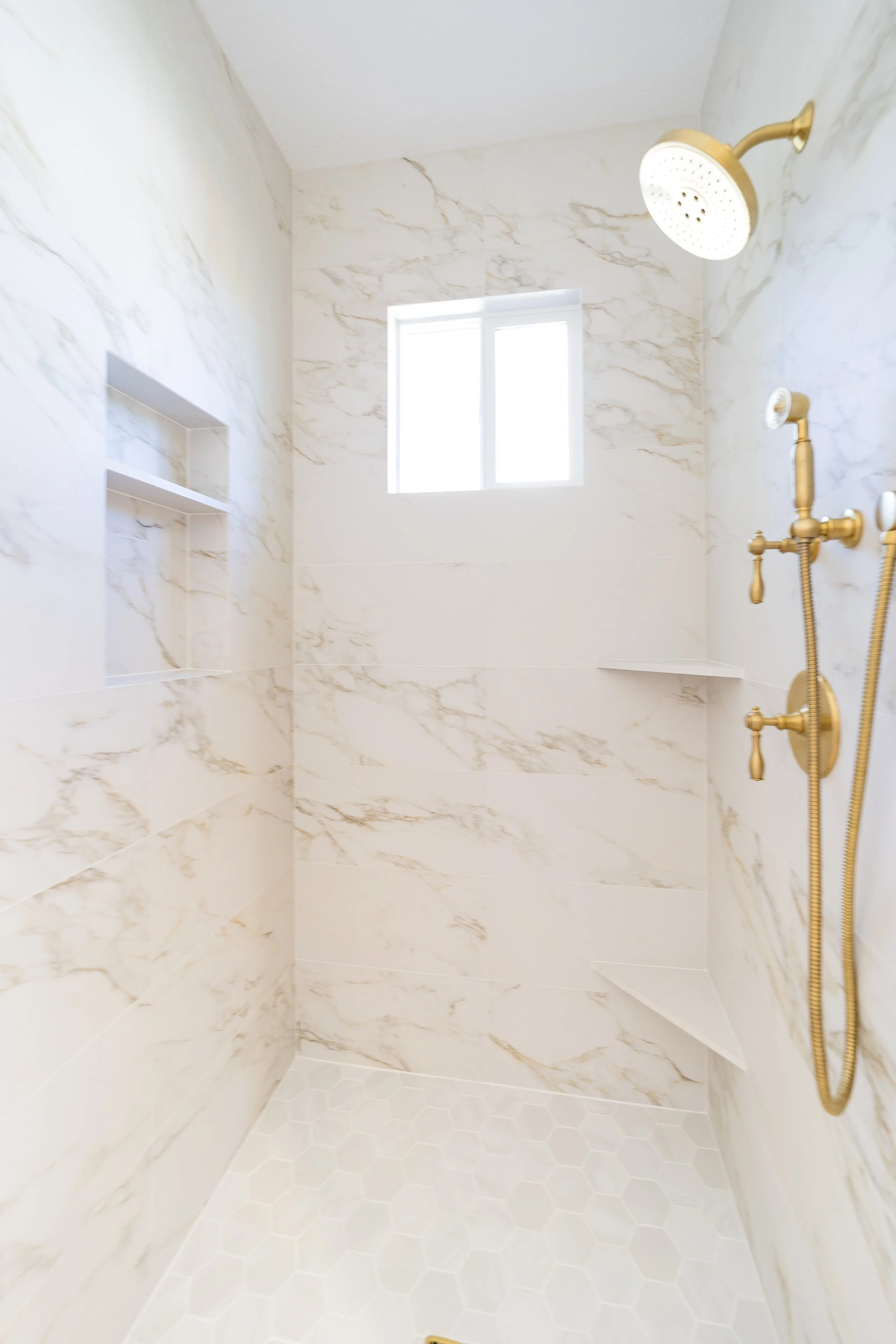 Shower with marble walls, small hexagon white floor tiles, gold fixtures, built-in shelves, and a small window.
