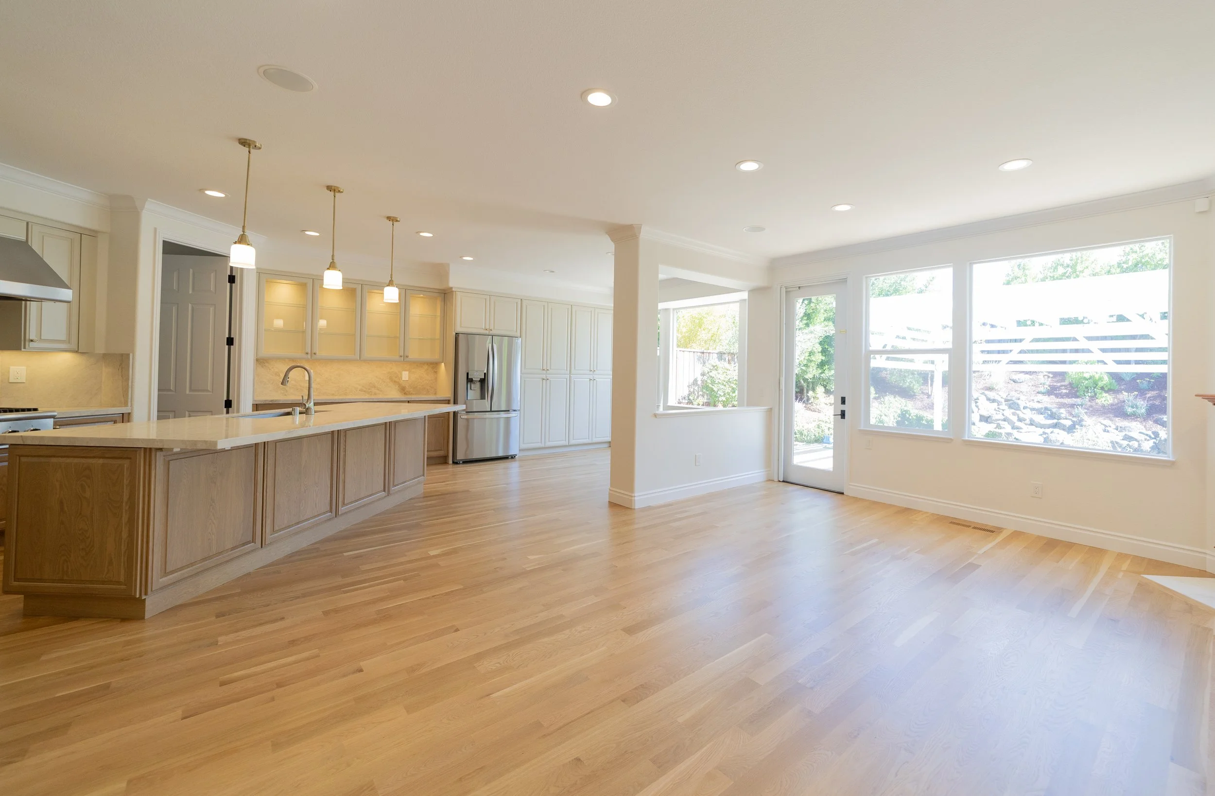 Empty kitchen and living area with hardwood floors, large windows, and a kitchen island with pendant lights.