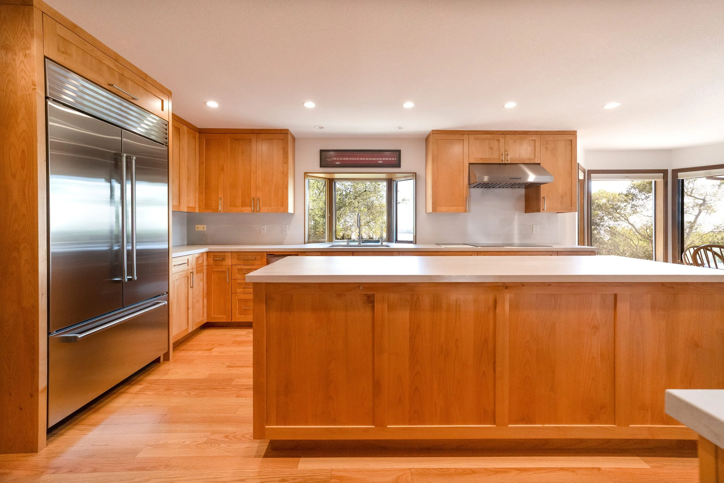 Modern kitchen with wooden cabinets, stainless steel refrigerator, large white island, and windows showing trees outside.