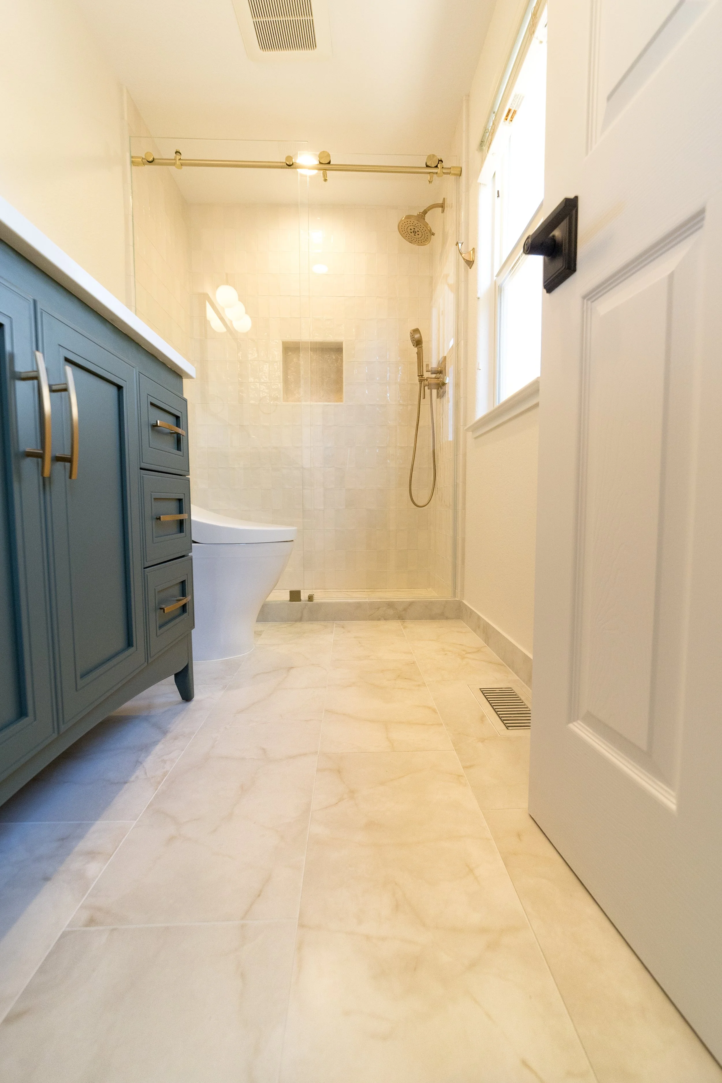 A bathroom with a tiled floor, a white toilet, a blue vanity cabinet, a glass shower with a wall niche, and a window letting in natural light.