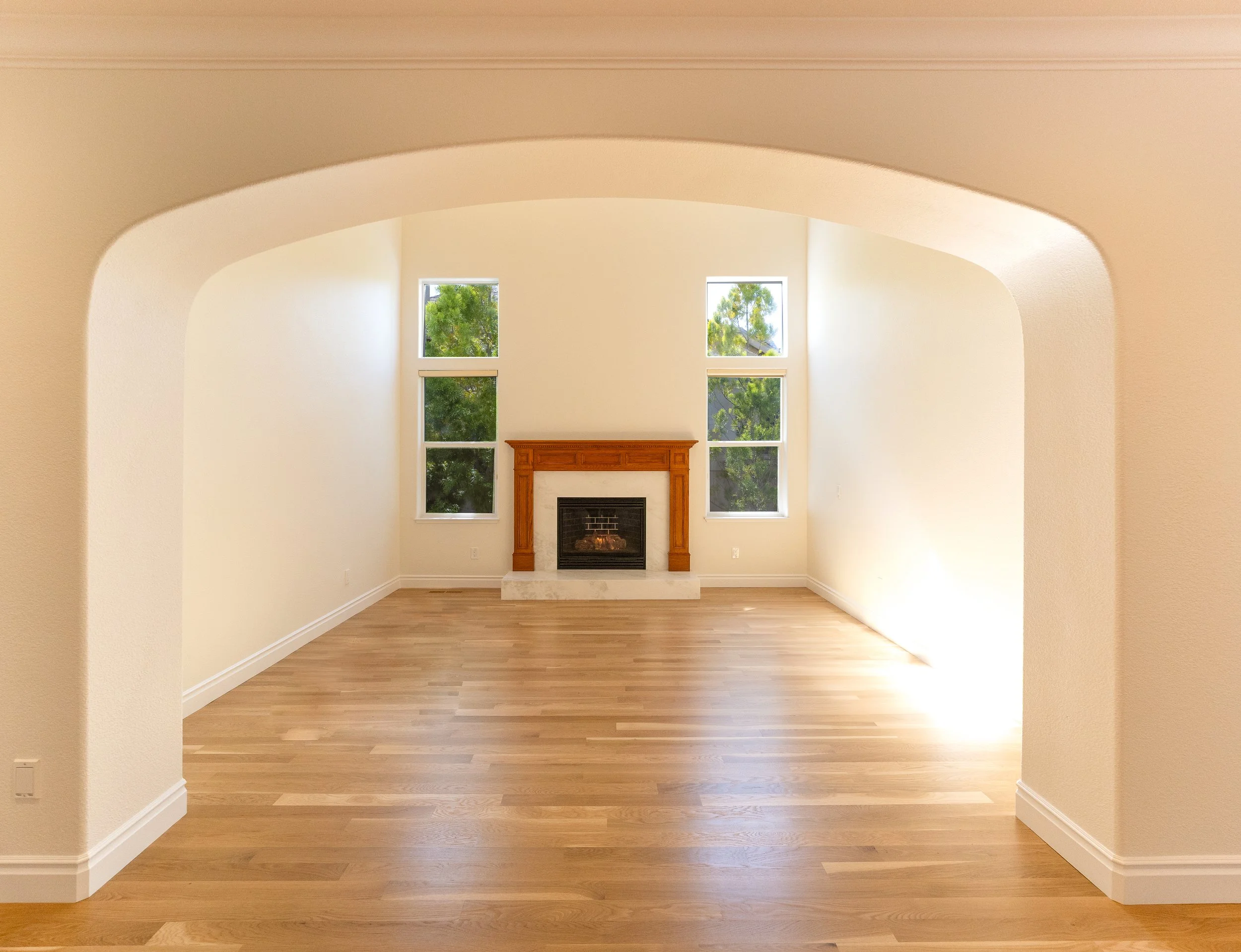 Empty living room with a fireplace, four windows, hardwood floor, and neutral-colored walls.