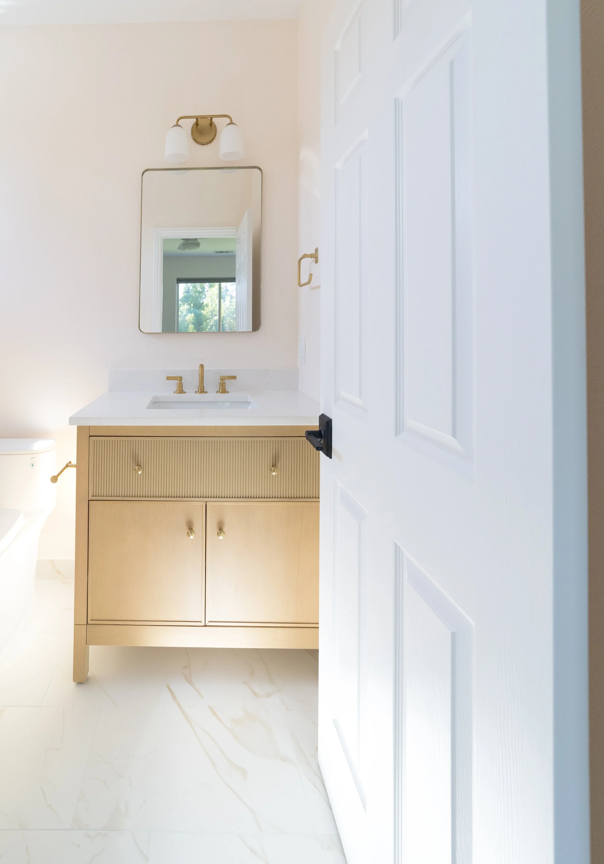 A partial view of a bathroom with a beige vanity, white countertop, gold fixtures, a mirror, and a partially opened white door.