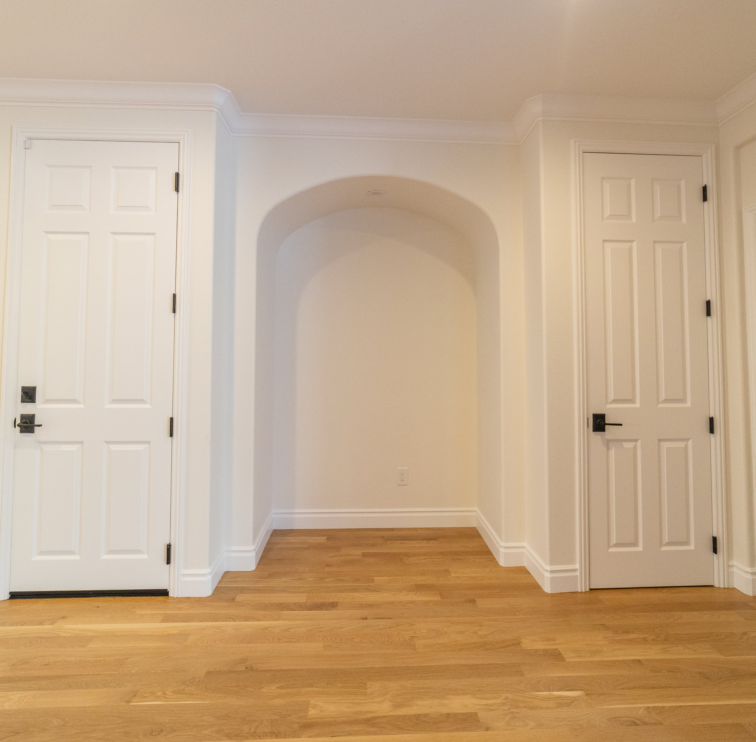 Empty room with white walls, three doors with black handles, and a wooden floor.