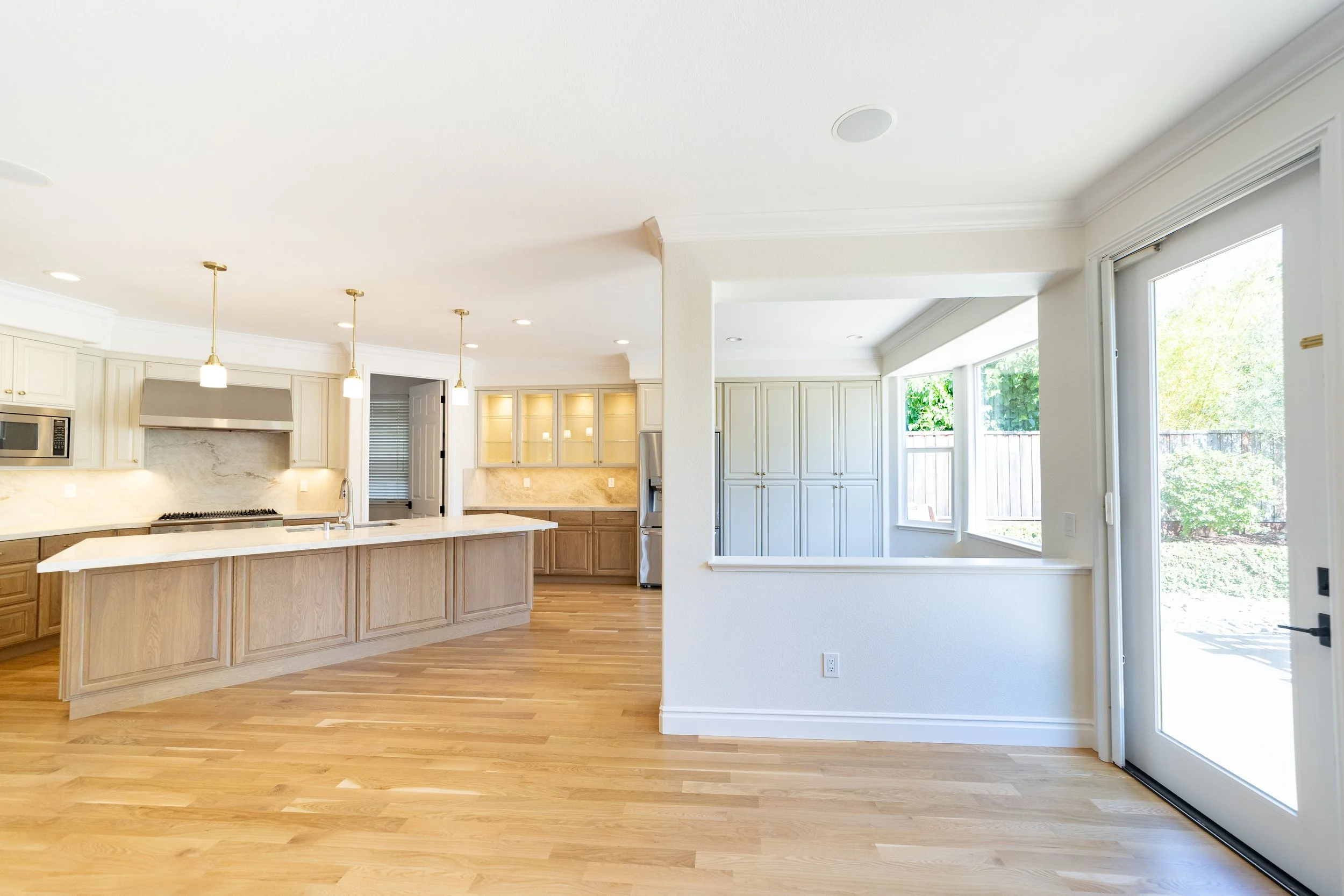 Bright open-concept kitchen with light wood cabinets, a large island, pendant lighting, and a view of a backyard through windows and a sliding glass door.