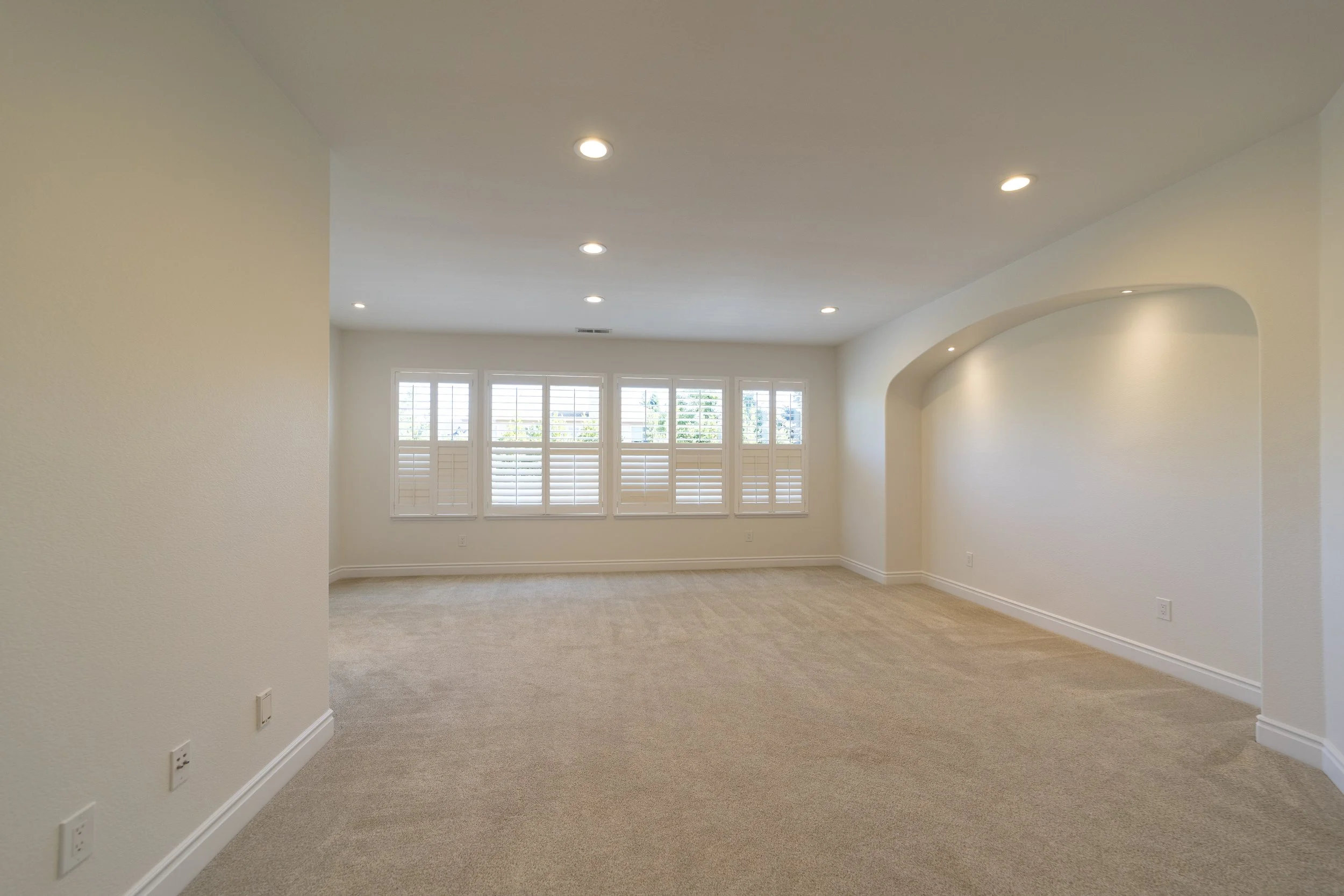Empty room with beige carpet flooring, white walls, large window with shutters, and recessed ceiling lights.