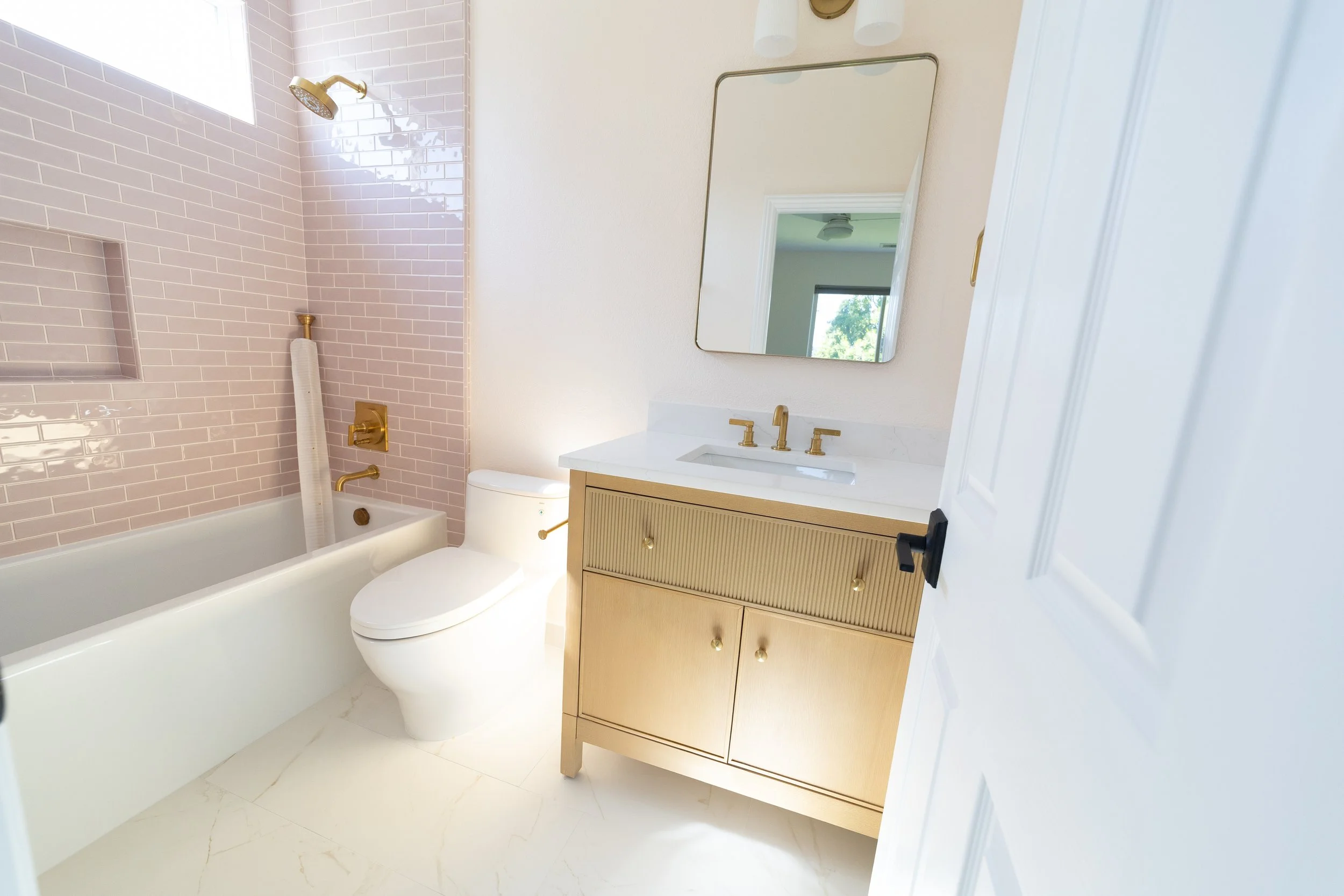 Bathroom with pink tiled shower wall, white bathtub, toilet, beige vanity with gold hardware, and a mirror