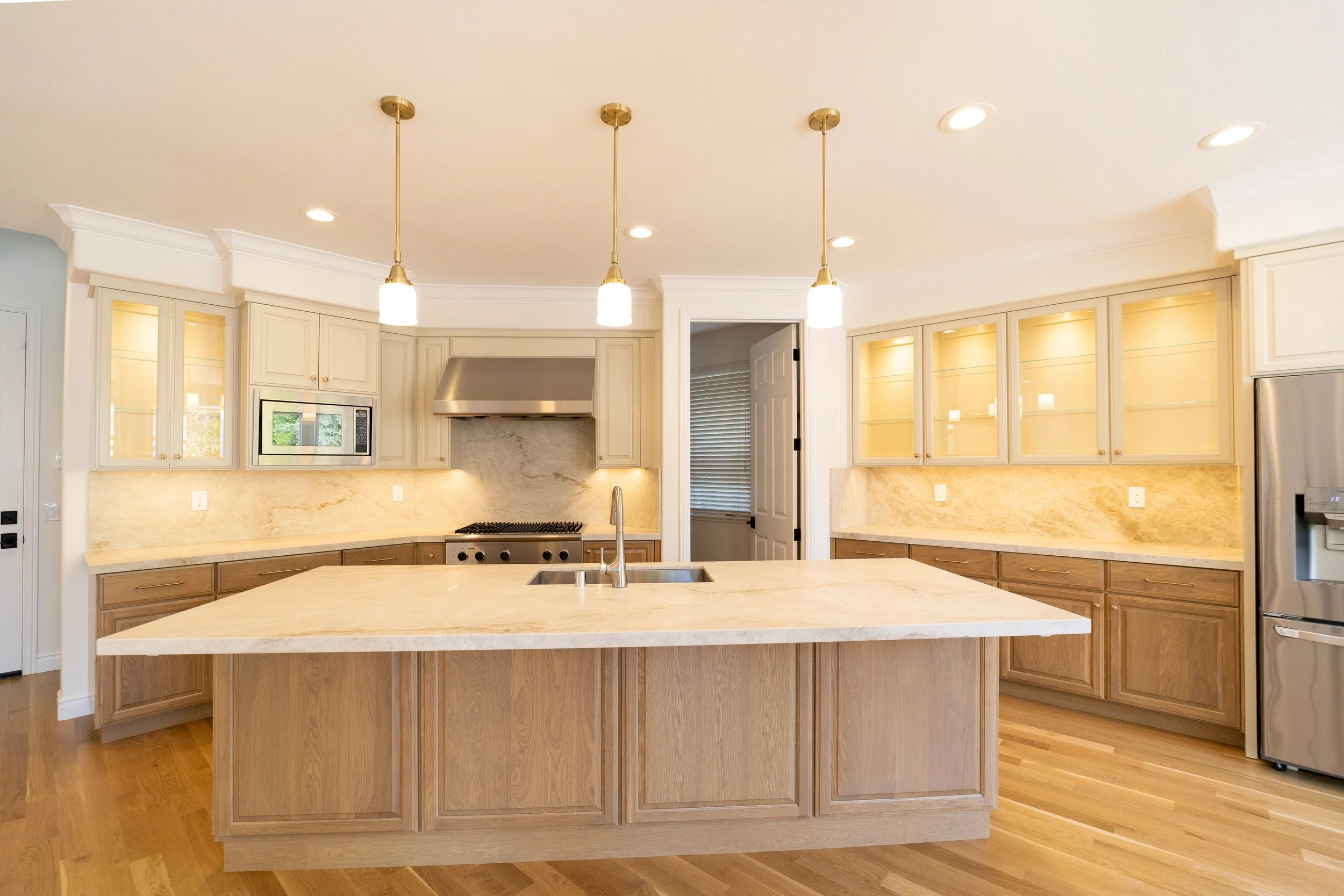 Modern kitchen with wooden cabinets, marble countertops, pendant lights, and stainless steel appliances, including a refrigerator and built-in microwave.