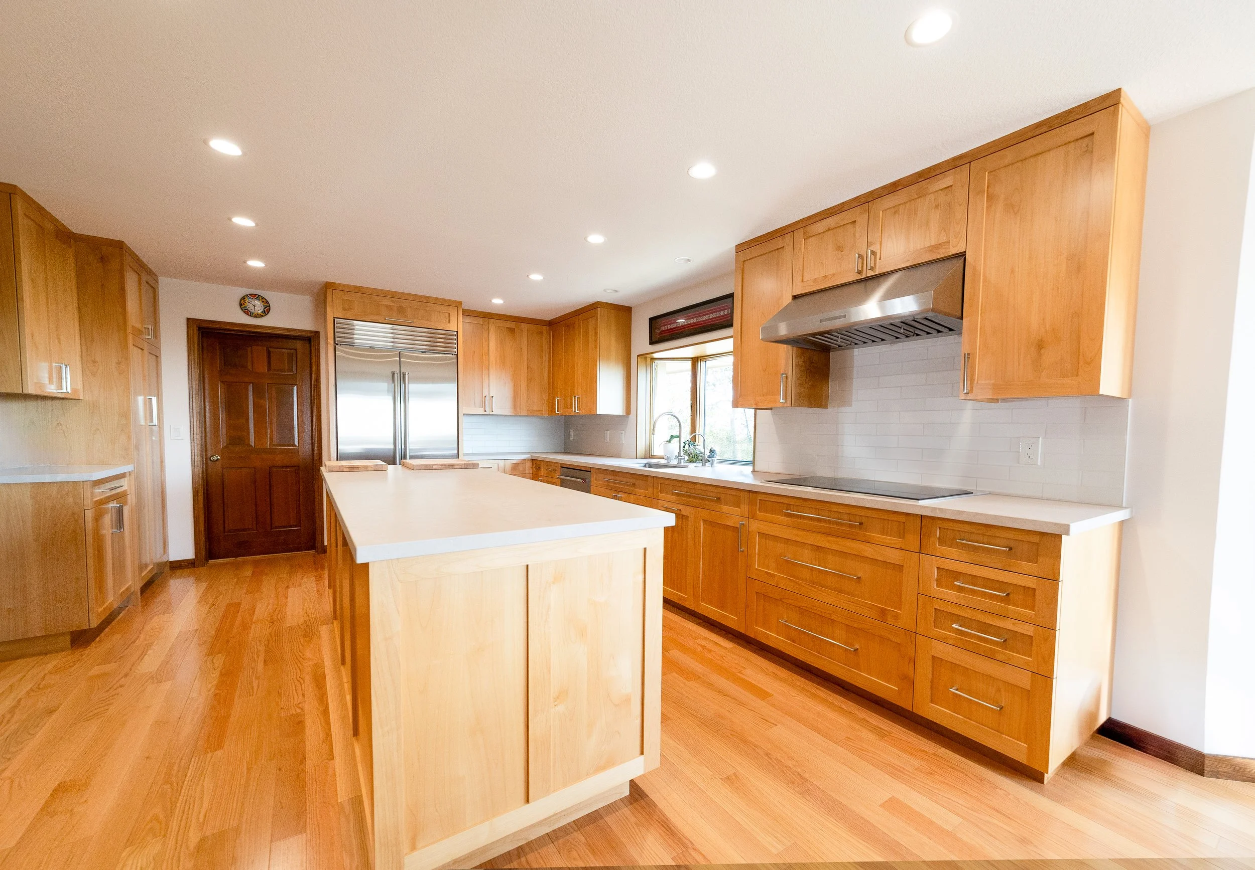 A spacious kitchen with wooden cabinets, a central island, a stainless steel refrigerator, a stove with a range hood, a window above the sink, and recessed ceiling lights.