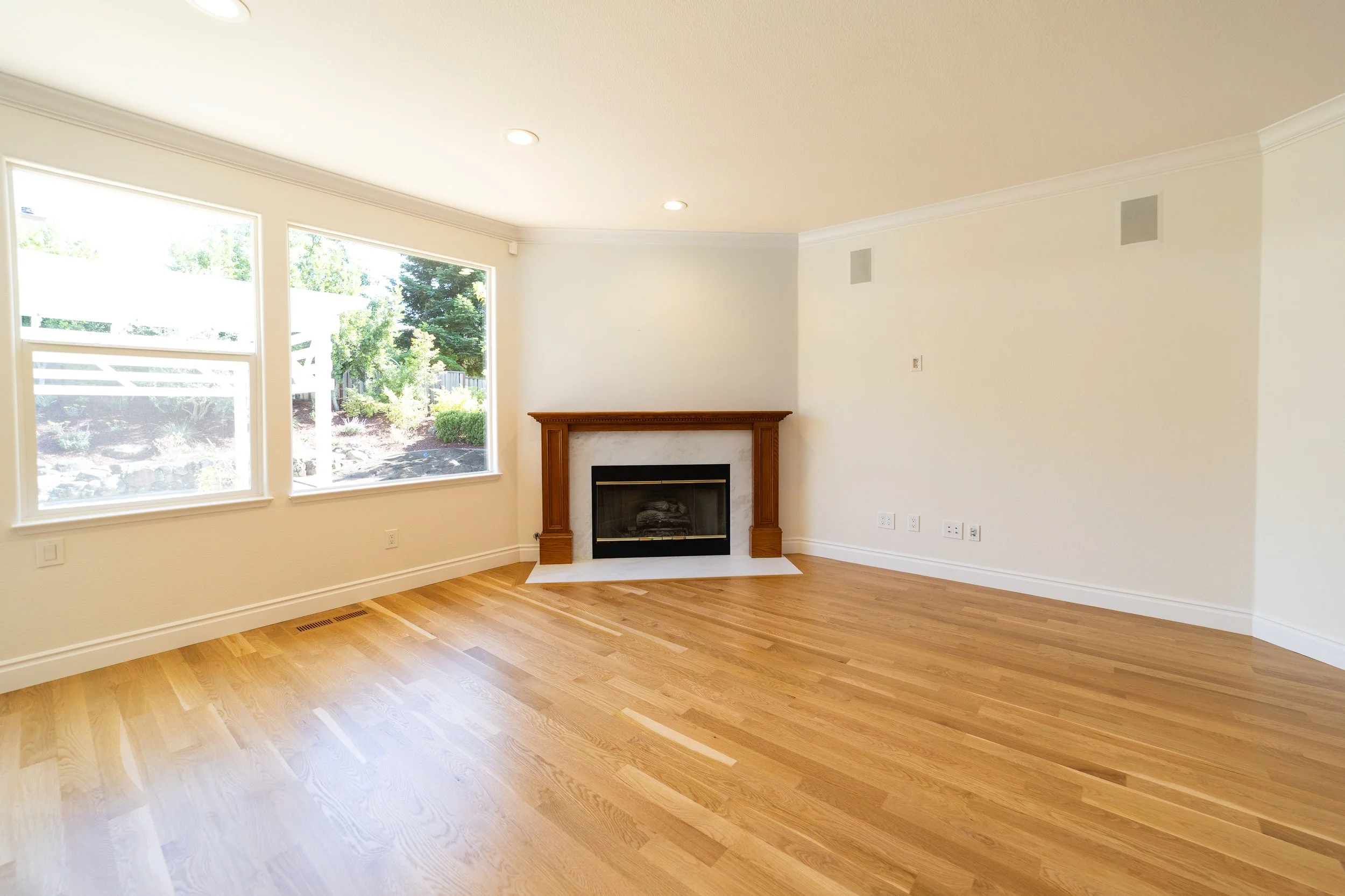Empty living room with hardwood floors, white walls, large windows, a fireplace with a wooden mantel, and recessed ceiling lights.