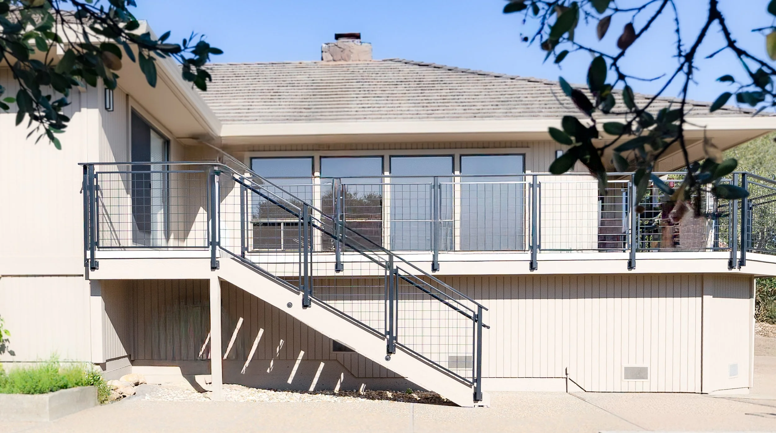 Modern house with a second-floor balcony and outdoor staircase, beige siding, large windows, and a chimney on the roof.