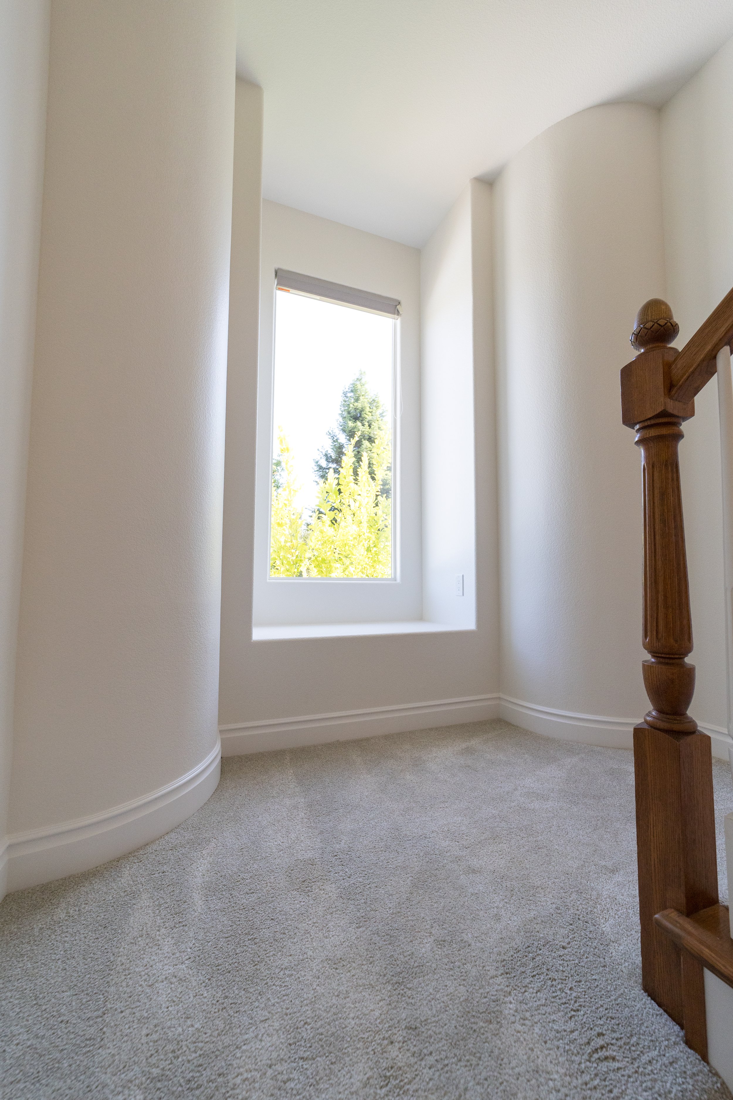 An empty hallway with beige carpet, white walls, a large window with a view of trees, and a wooden staircase railing.