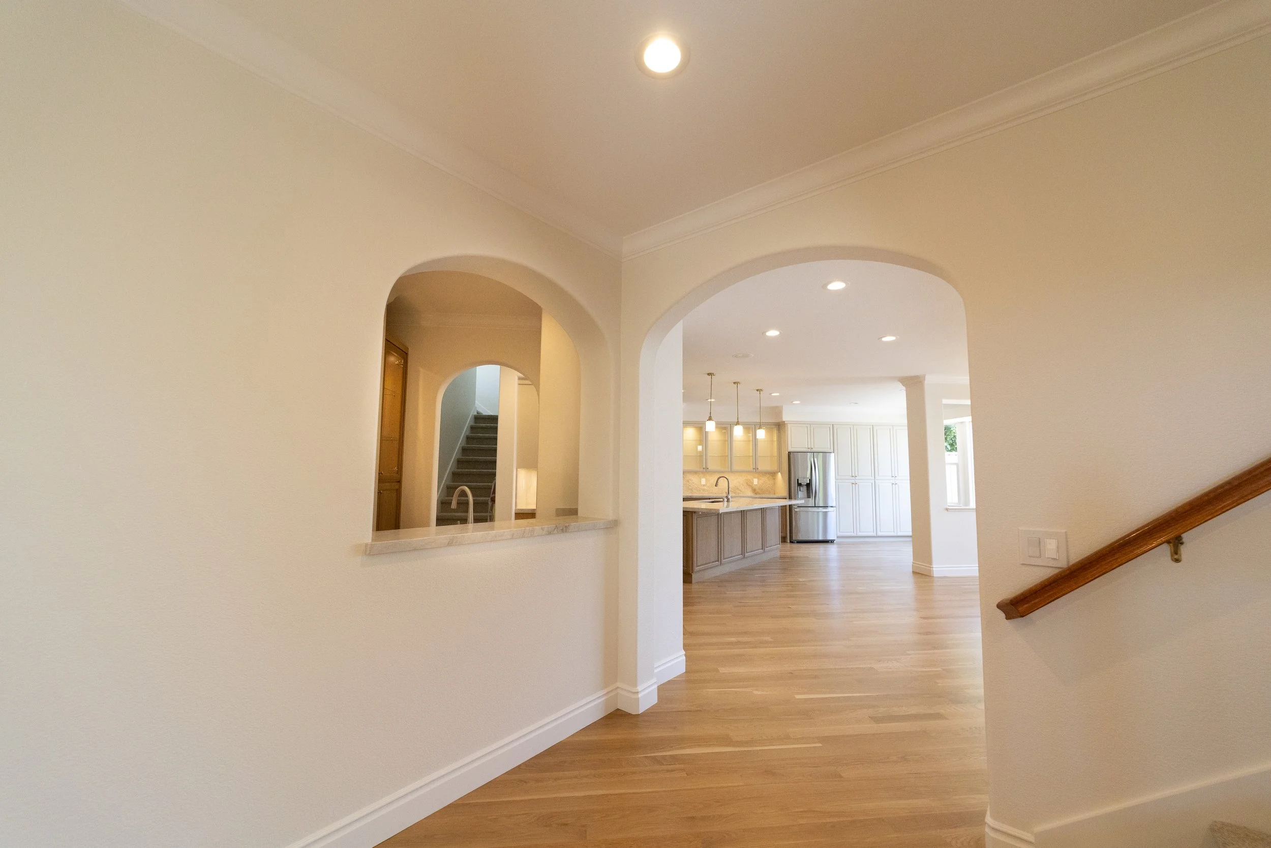 Interior view of a bright, spacious home with a kitchen in the background. The space has light-colored walls, wooden flooring, and arched openings leading to different areas. Recessed lighting is visible on the ceiling.