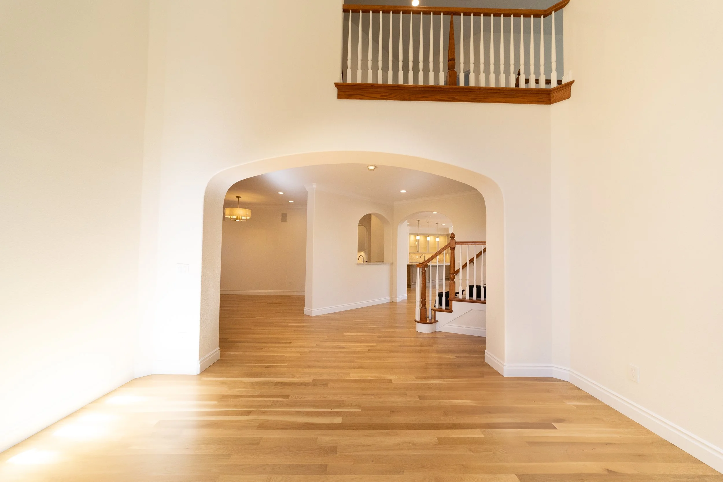 Empty living room with hardwood floors, white walls, arched doorways, and a staircase with wooden railing leading to an upper level.