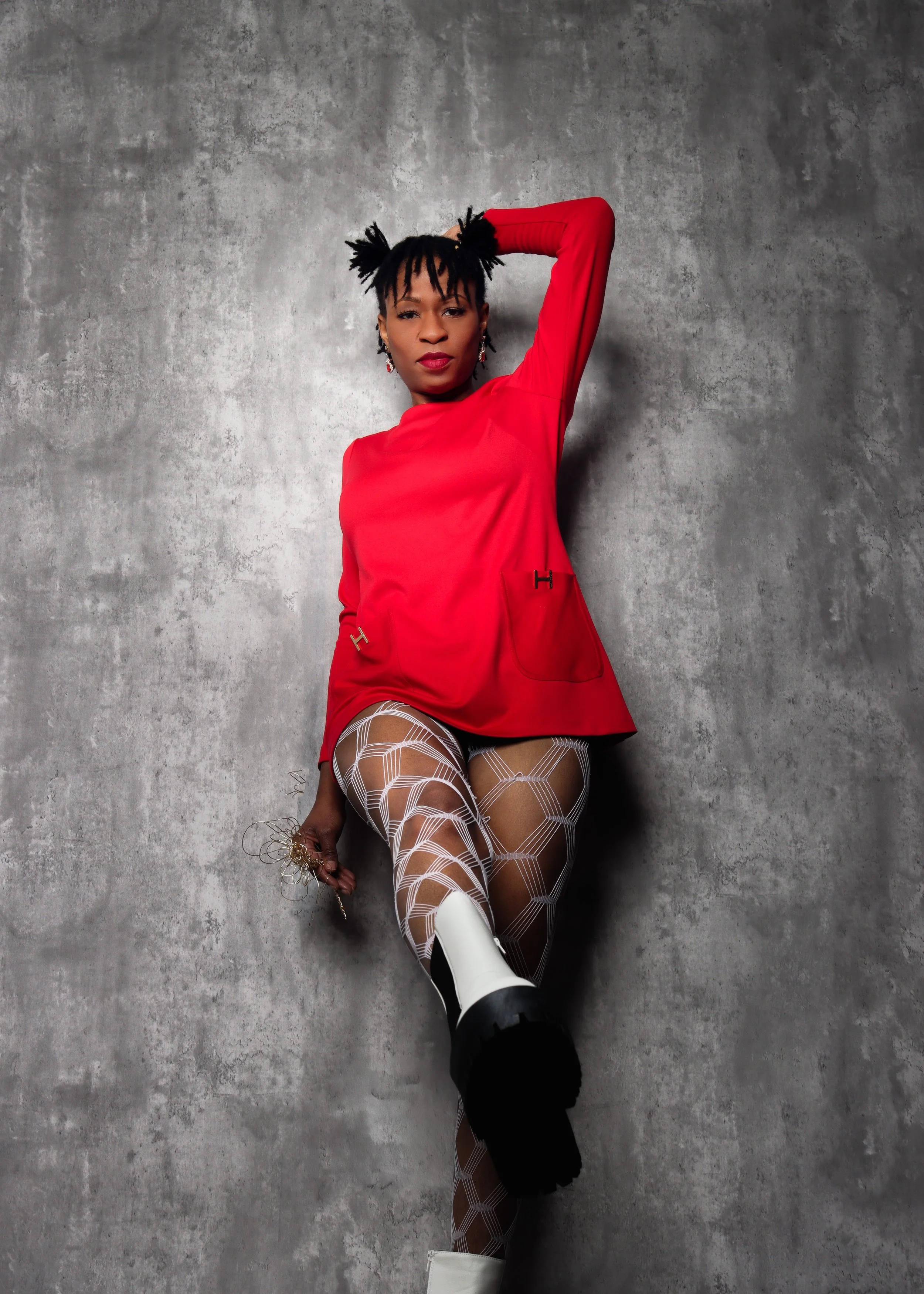 Person in a red top and patterned tights posing against a concrete wall.