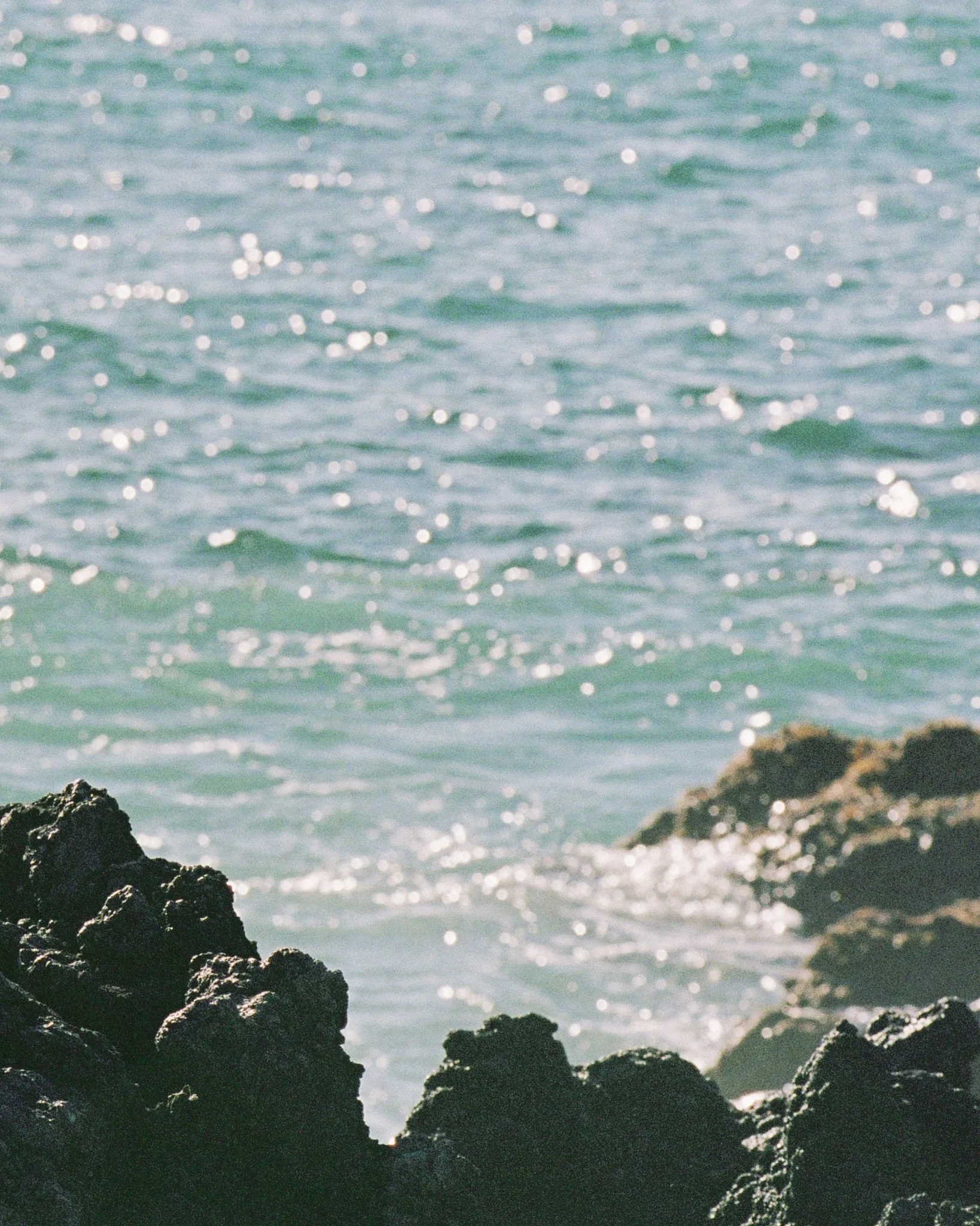 Rocks on a seashore with waves and sunlight reflecting on the water.