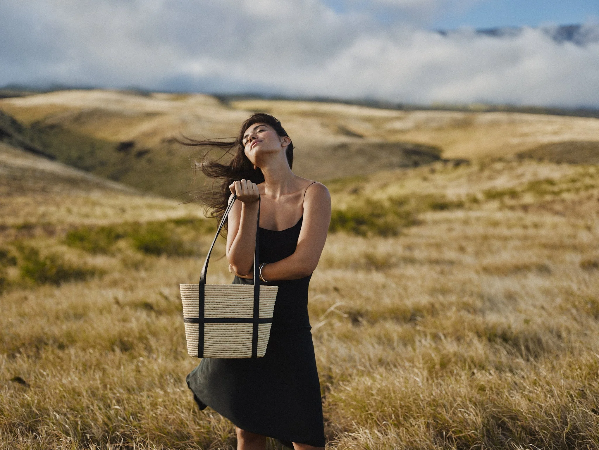 A woman in a black dress holding a beige tote bag with black handles stands in a grassy field with rolling hills in the background during daytime.