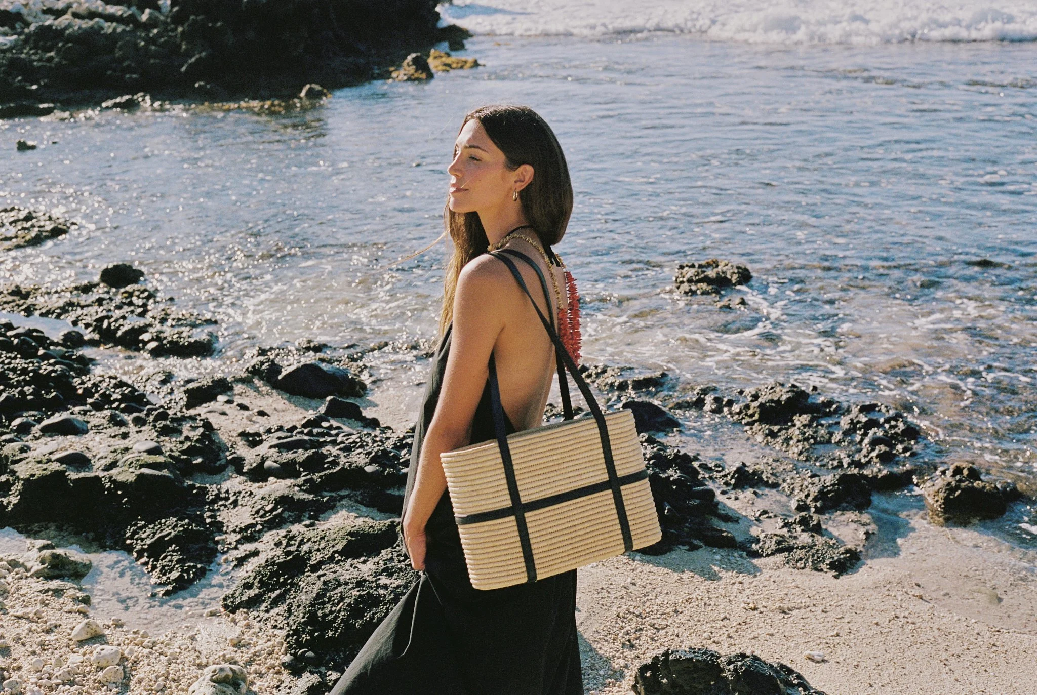 A woman with long dark hair standing on a rocky beach with ocean waves in the background, carrying a beige and black striped handbag over her shoulder.