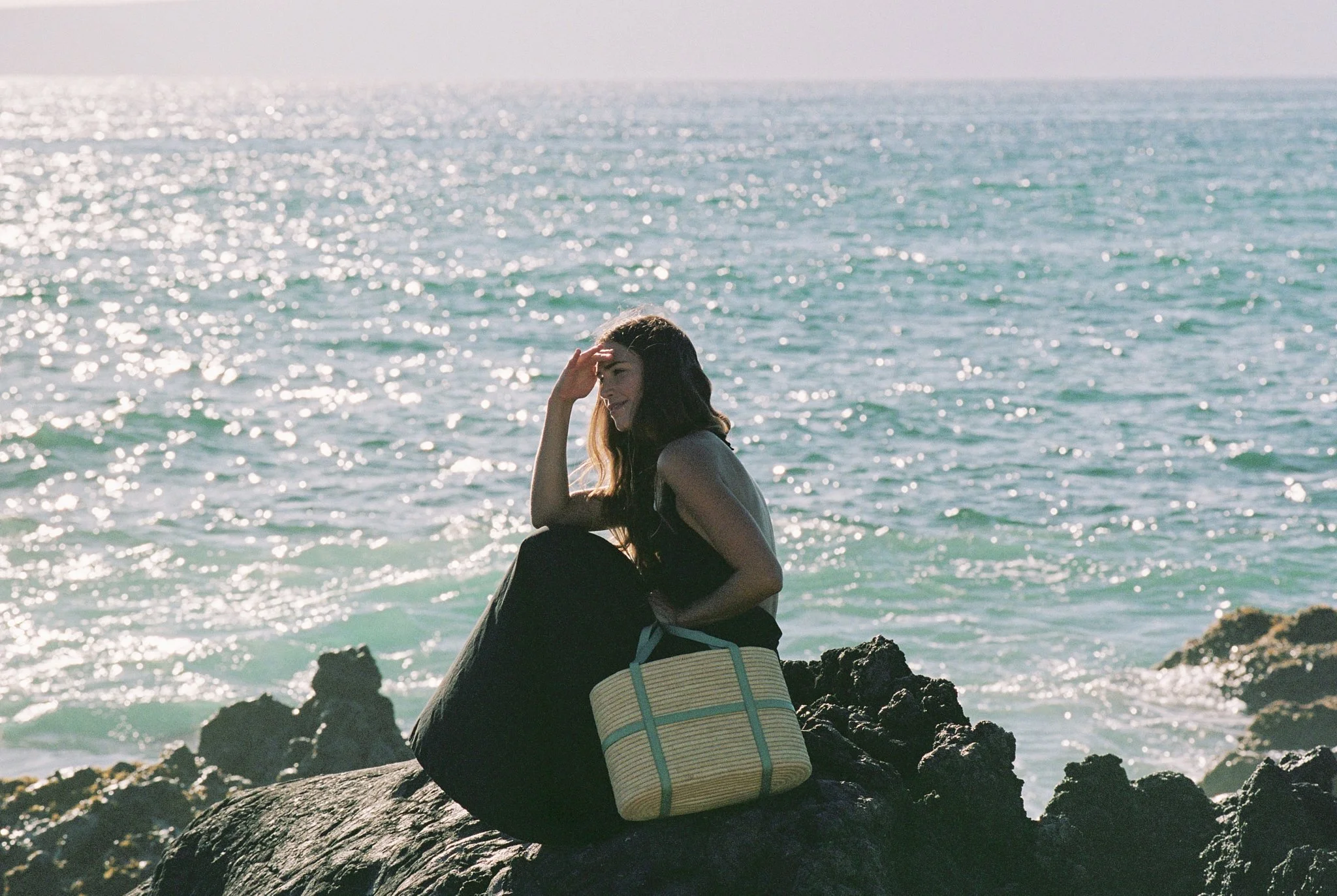 A woman sitting on rocks by the ocean, wearing a black dress, with a striped bag next to her, shielding her eyes from the sun, during sunset.