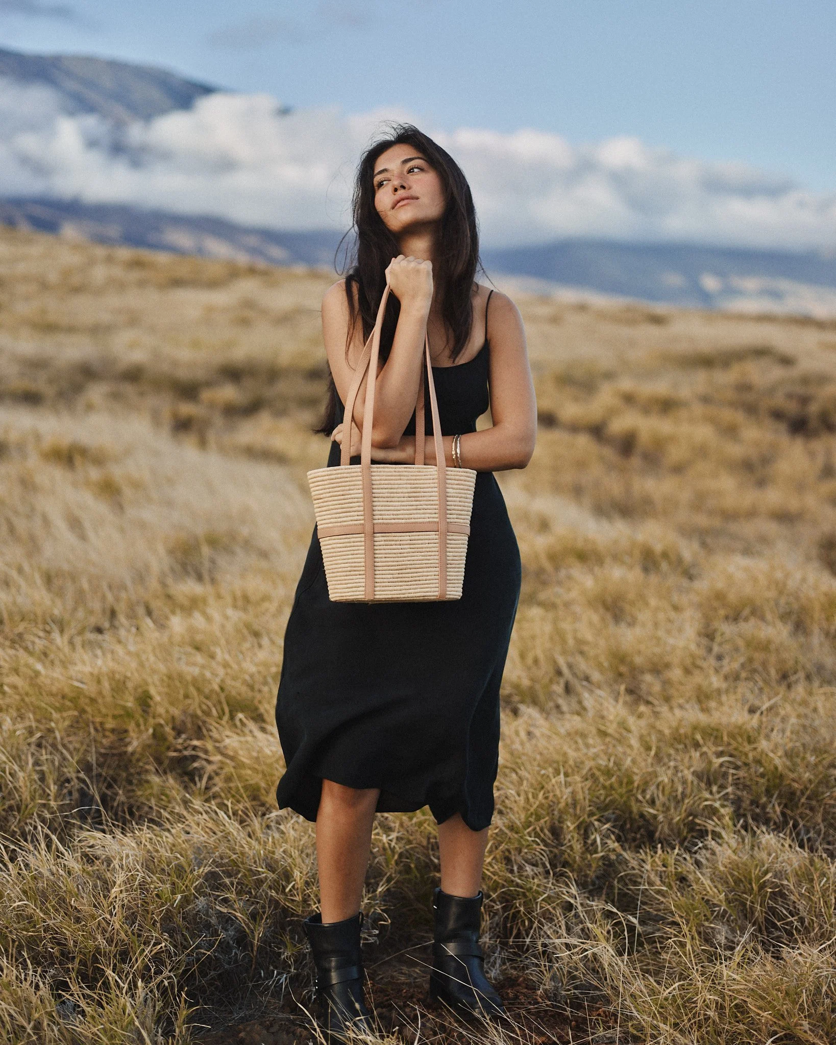 A woman in a black dress and black boots carrying a beige woven tote bag stands in a grassy field with mountains and cloudy sky in the background.