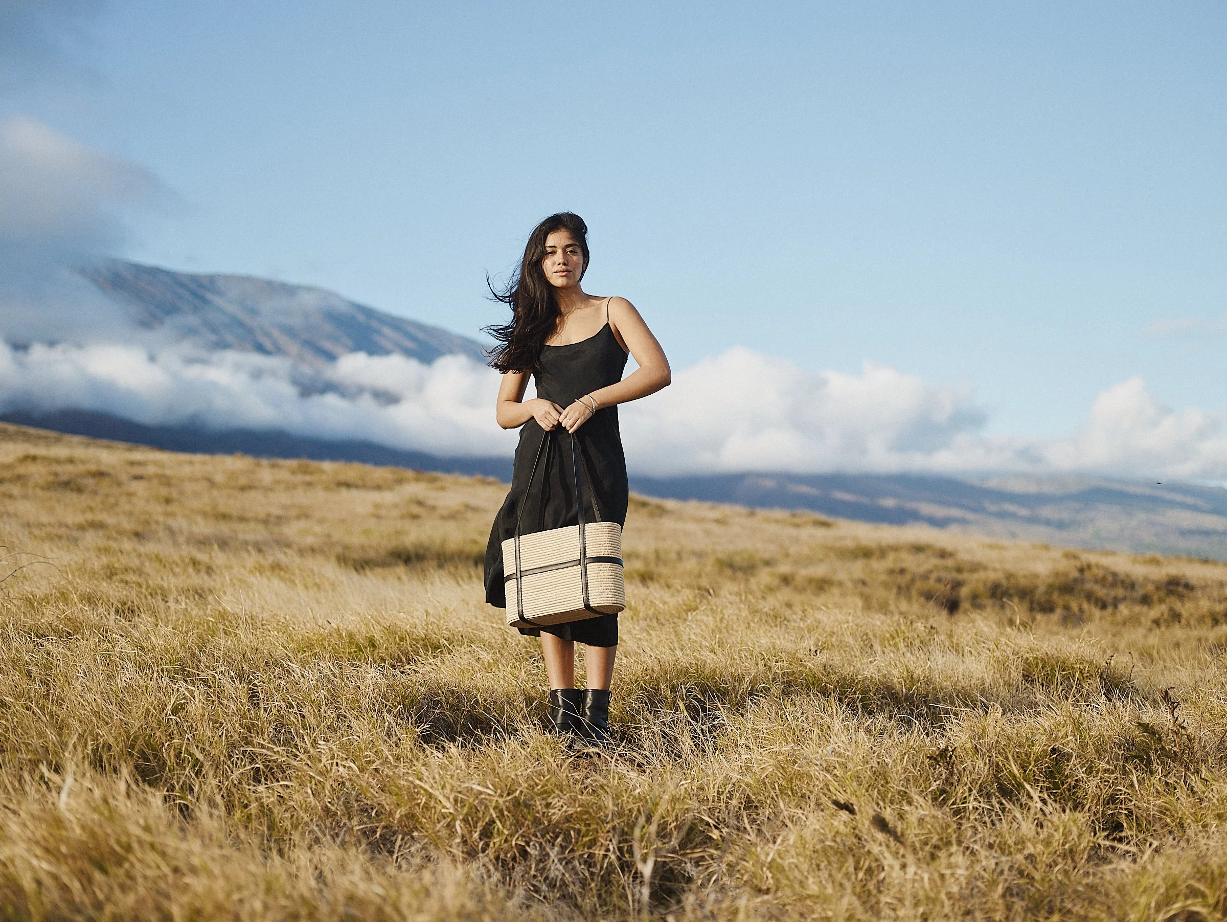 A young woman in a black dress standing in a grassy field with mountains and partly cloudy sky in the background, holding a striped tote bag.