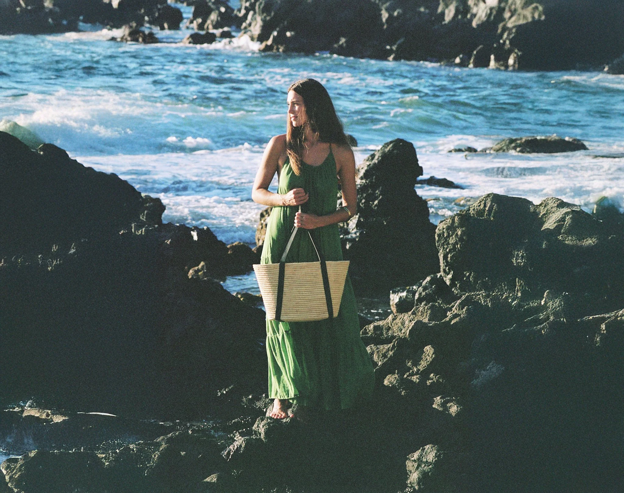A woman in a long green dress standing on rocks by the ocean, holding a striped tote bag, with waves and rocky shoreline in the background.