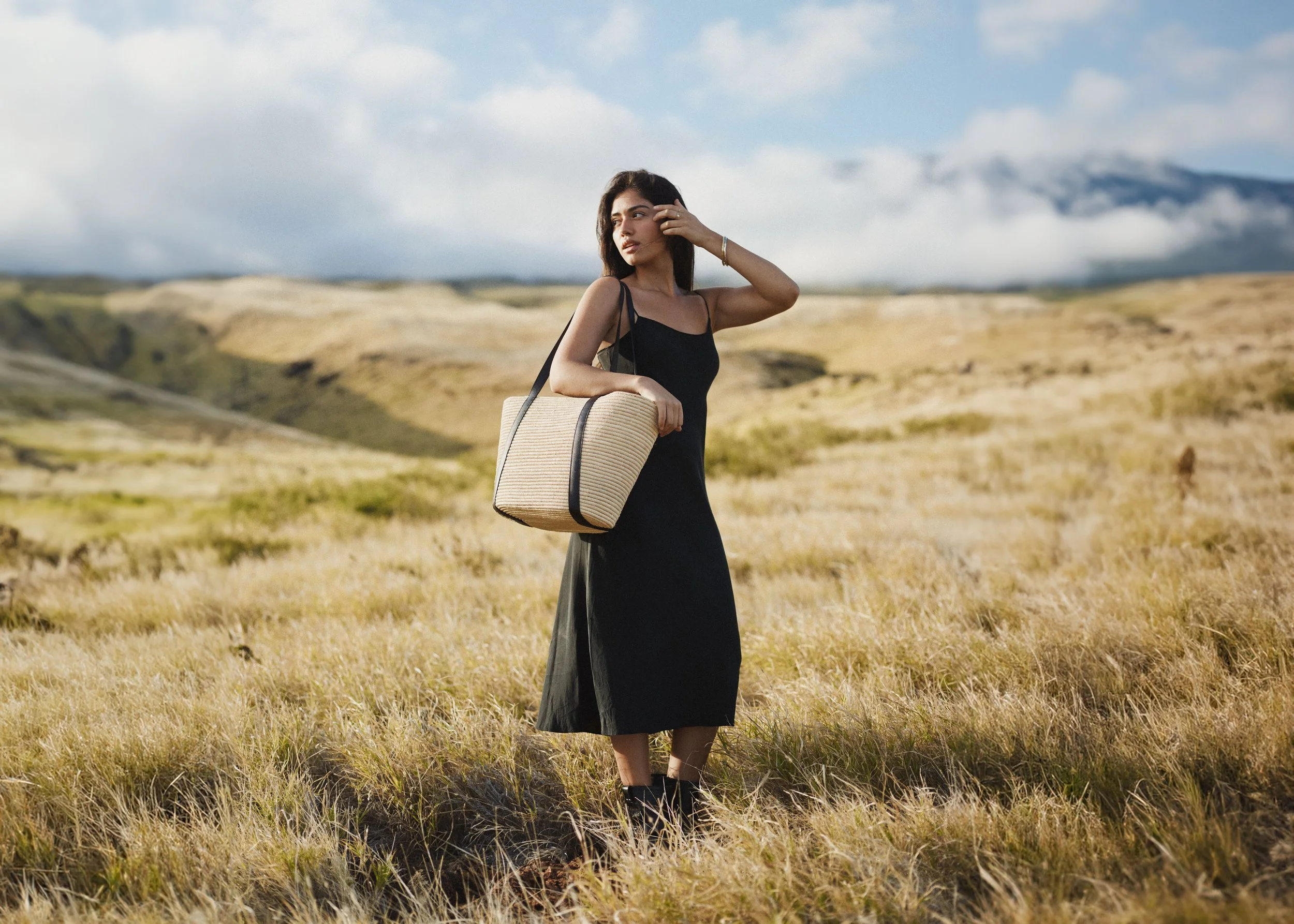 A woman in a black dress standing in a grassy field, carrying a straw bag, with mountains and a cloudy sky in the background.