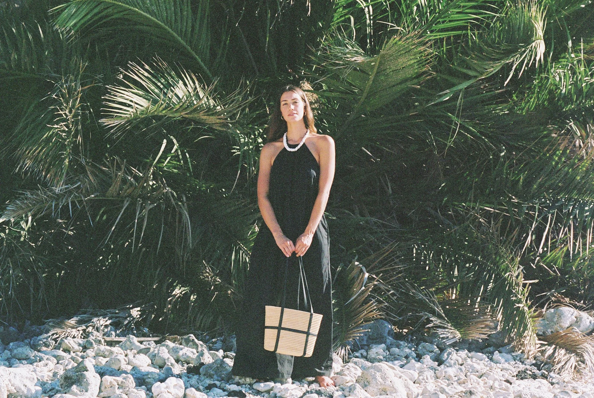 A woman in a black dress and necklace stands barefoot on a rocky beach, holding a woven tote bag, with dense green palm leaves in the background.
