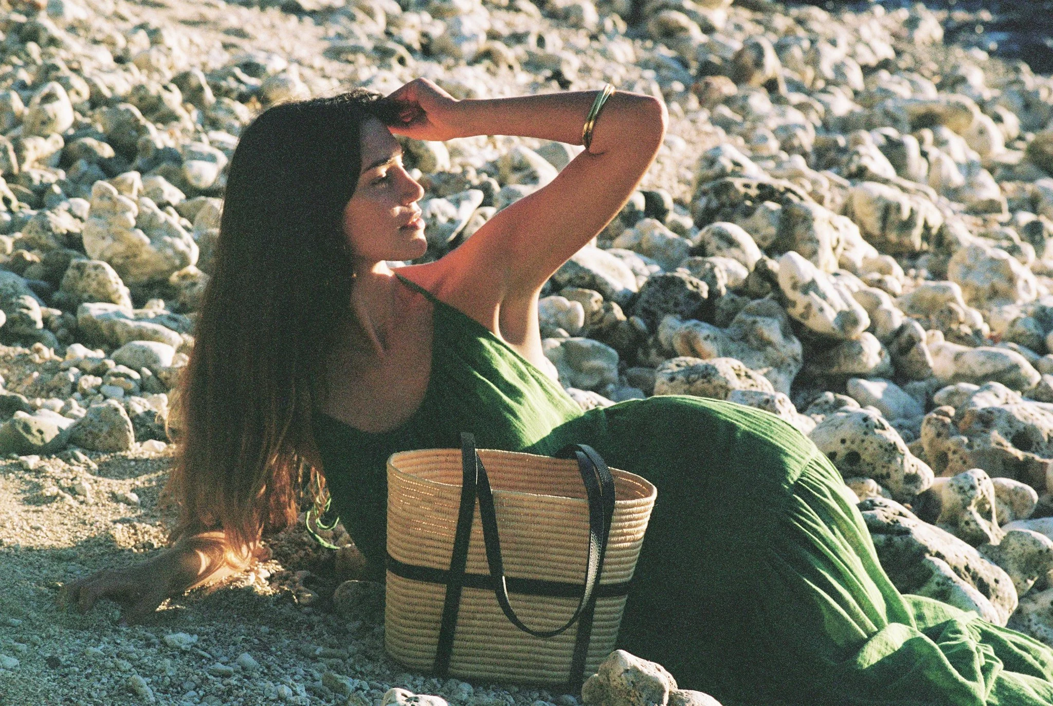 A woman in a green dress relaxing on a rocky beach, lying on the sand with stones, with a woven handbag nearby.