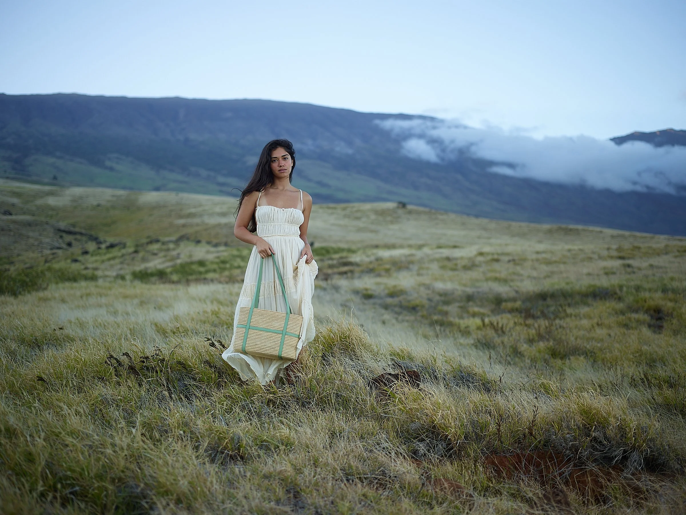 A woman in a white dress standing in a grassy field with mountains and clouds in the background, holding a woven tote bag.