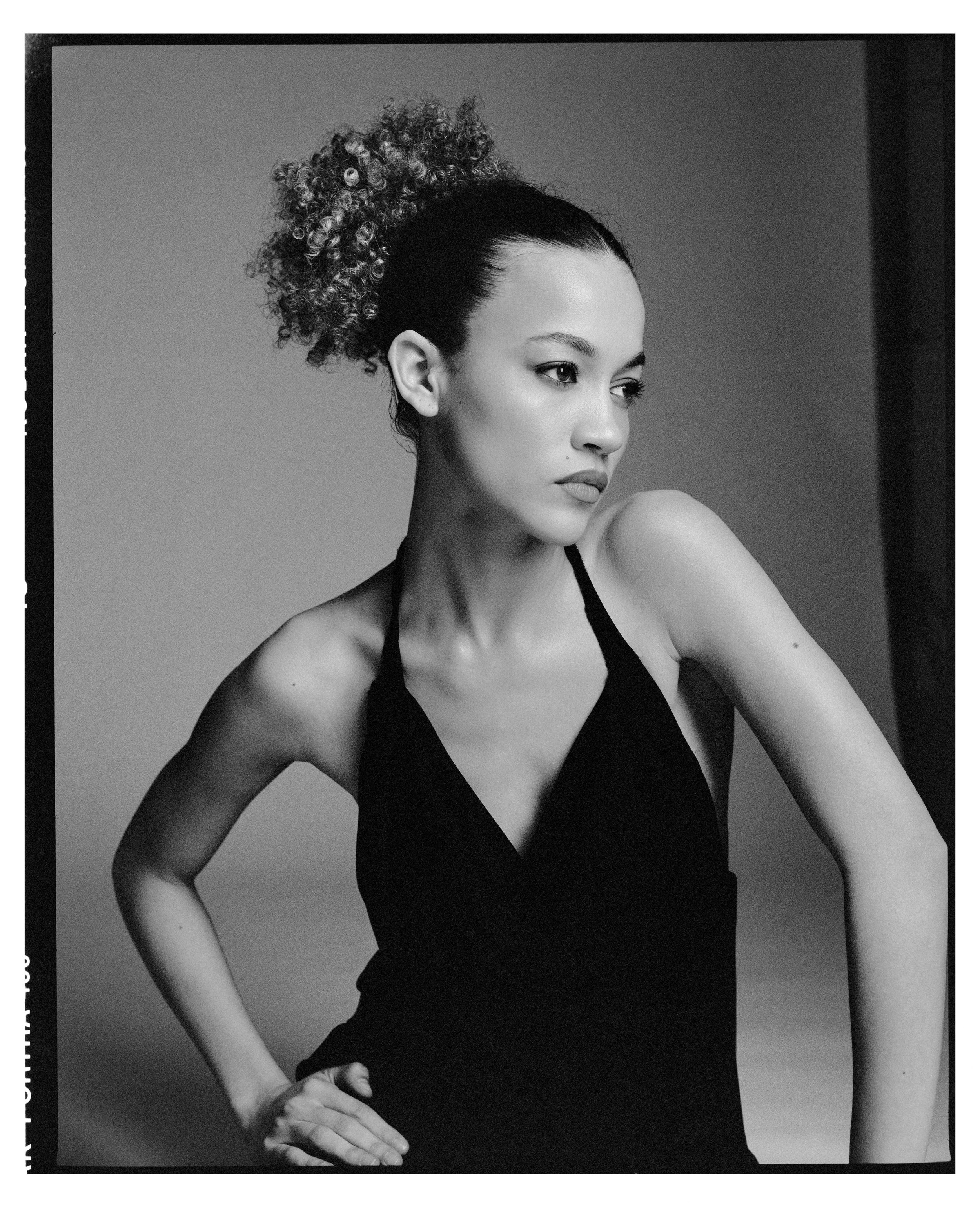 Black and white portrait of a woman with curly hair styled in a high bun, wearing a sleeveless top, with her hand on her hip and looking to her left.