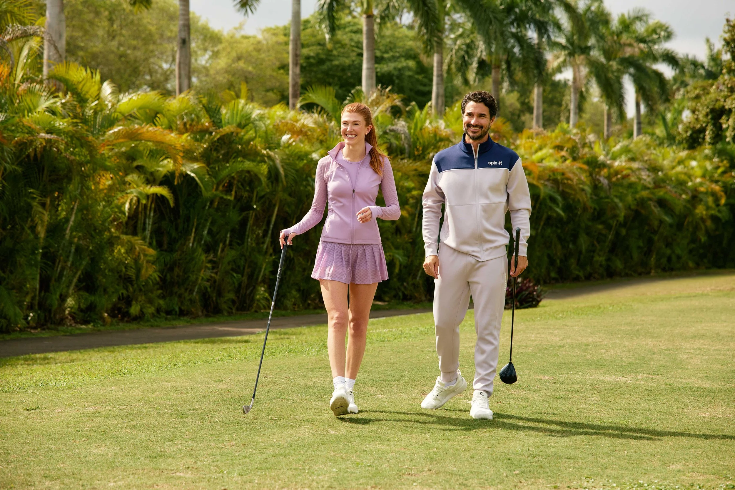A smiling woman and man walking with golf clubs on a golf course surrounded by trees and greenery.