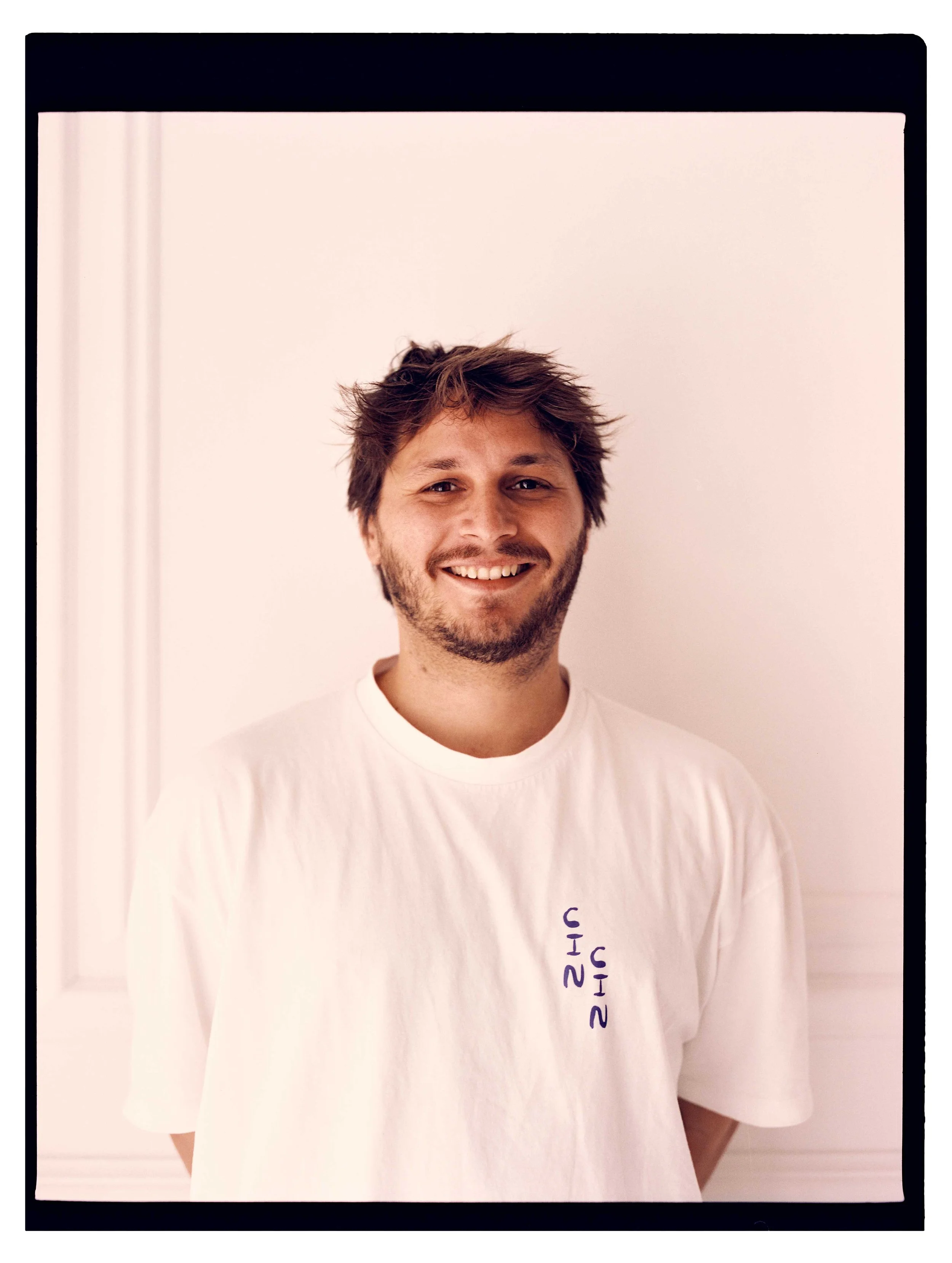 A smiling young man with tousled brown hair and a beard, wearing a white T-shirt with some text, standing against a plain white background.