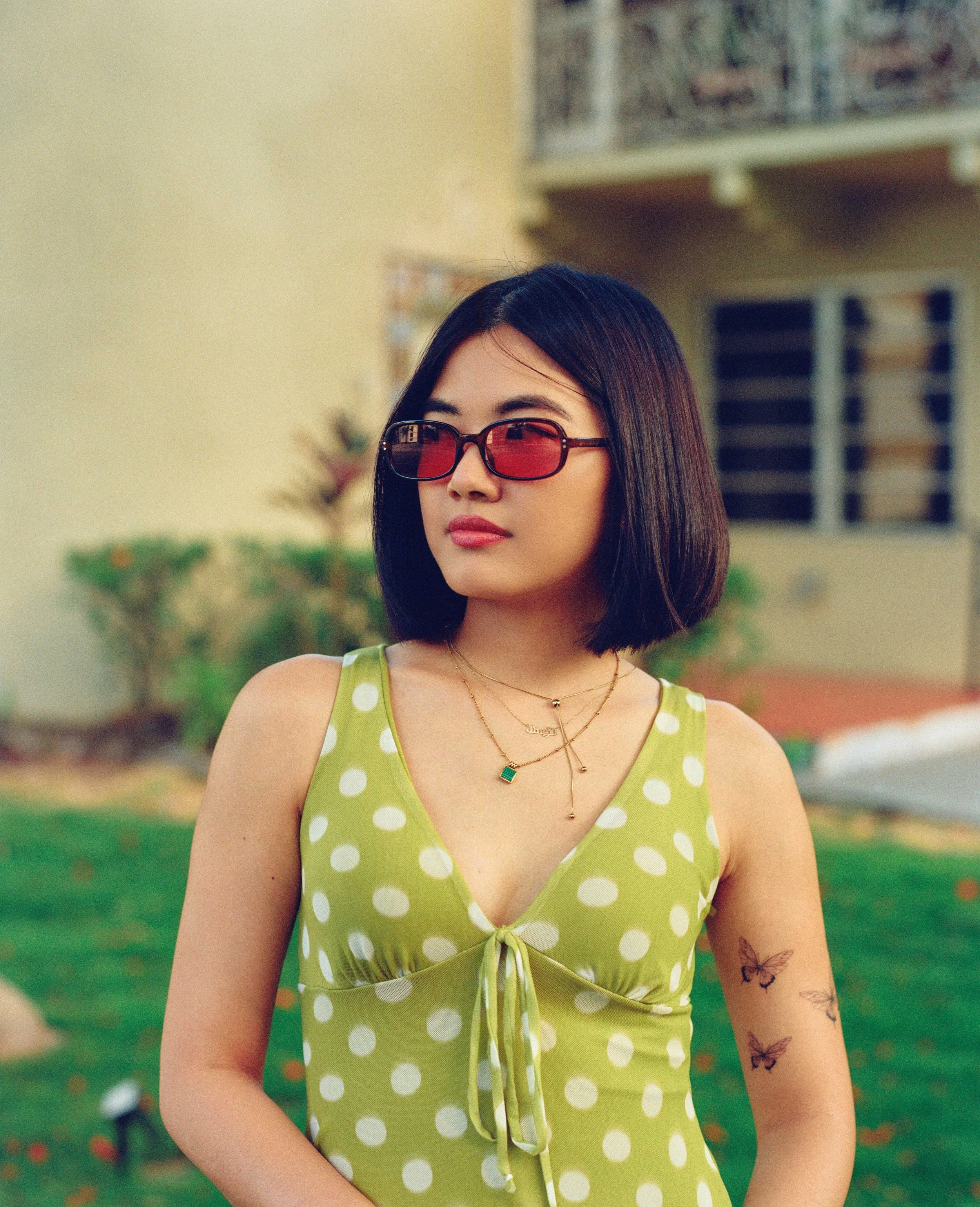 A woman with shoulder-length black hair wearing large square sunglasses with red lenses and a green sleeveless dress with white polka dots, standing outdoors in front of a house with windows and greenery.