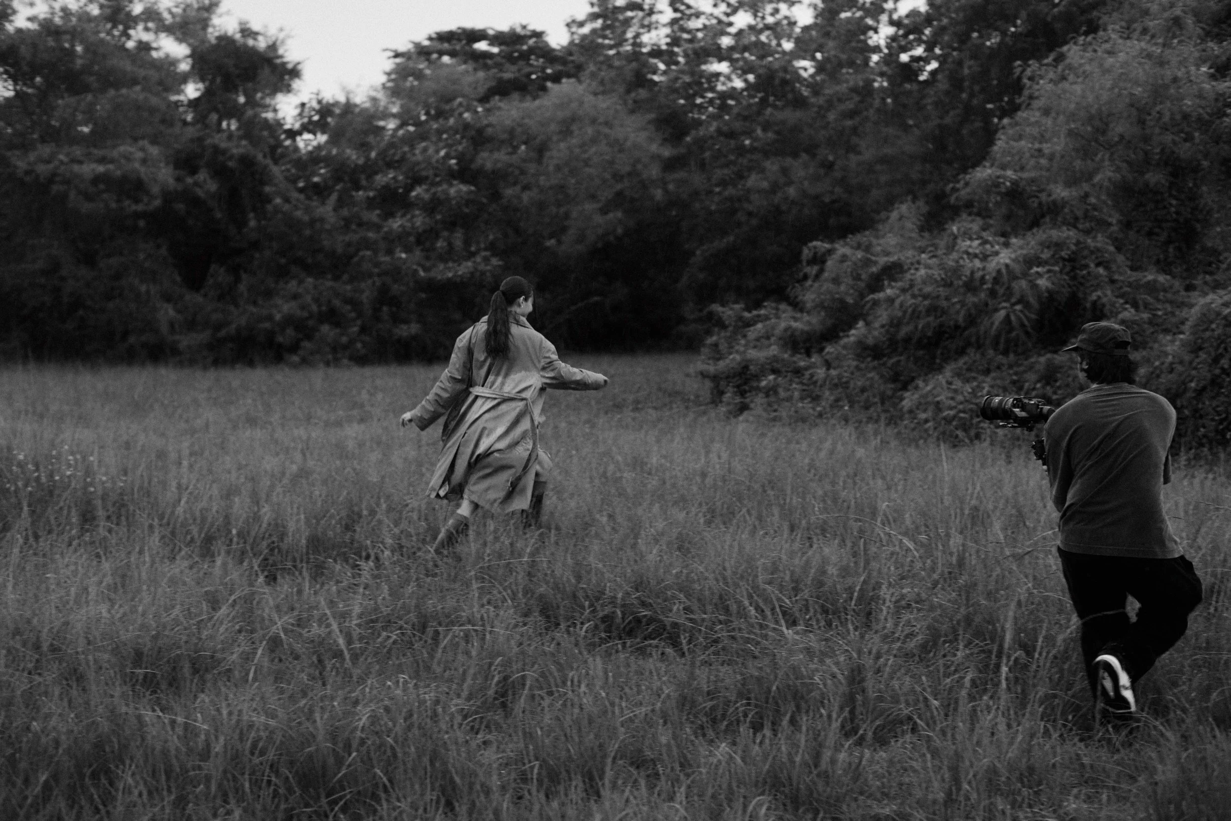 A woman in a coat running in a grassy field while a man with a camera crouches nearby, with trees in the background.
